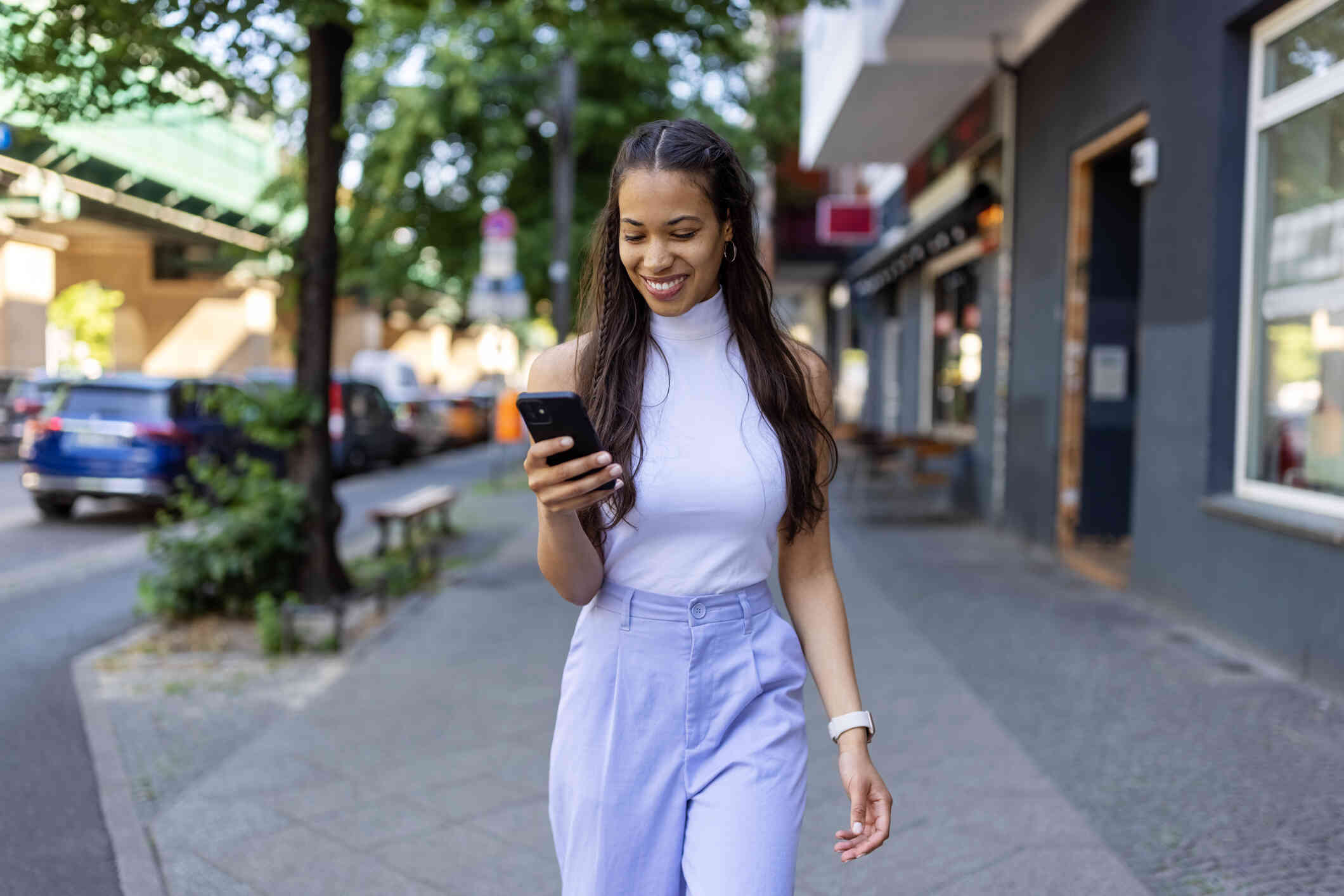 A woman in a white top walks outside down a sidwalk on a sunny day while smiling down at the cellphone in her hand.