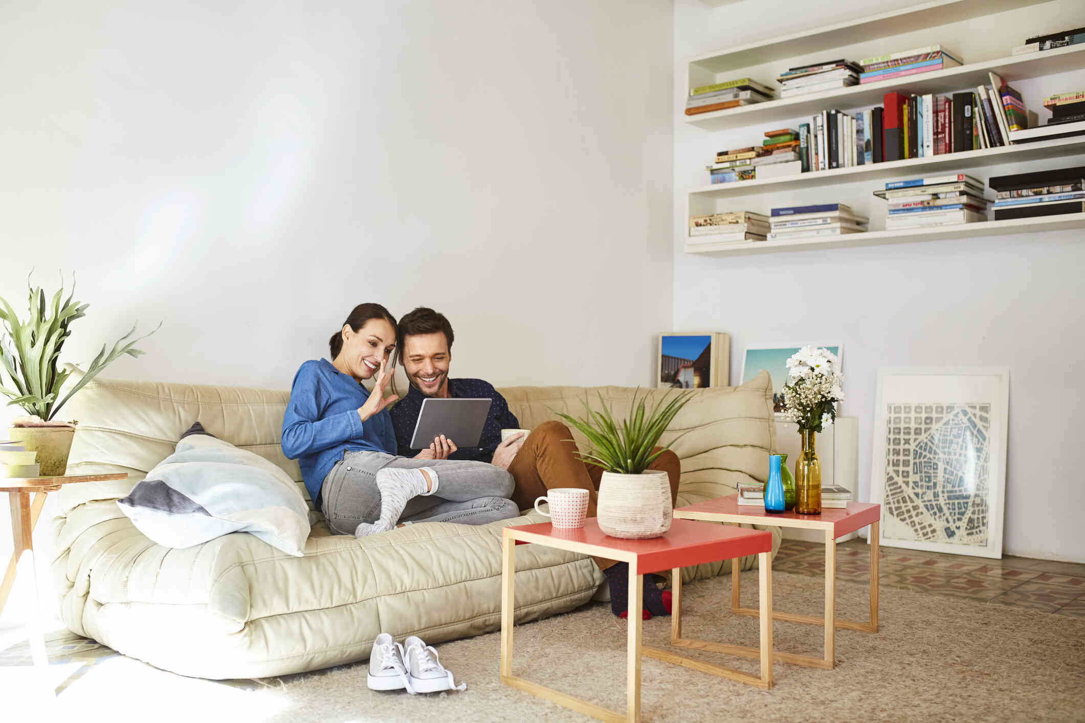 A man and a woman with dark brown hair smile as they sit on their couch and interact with someone on a tablet in front of them.