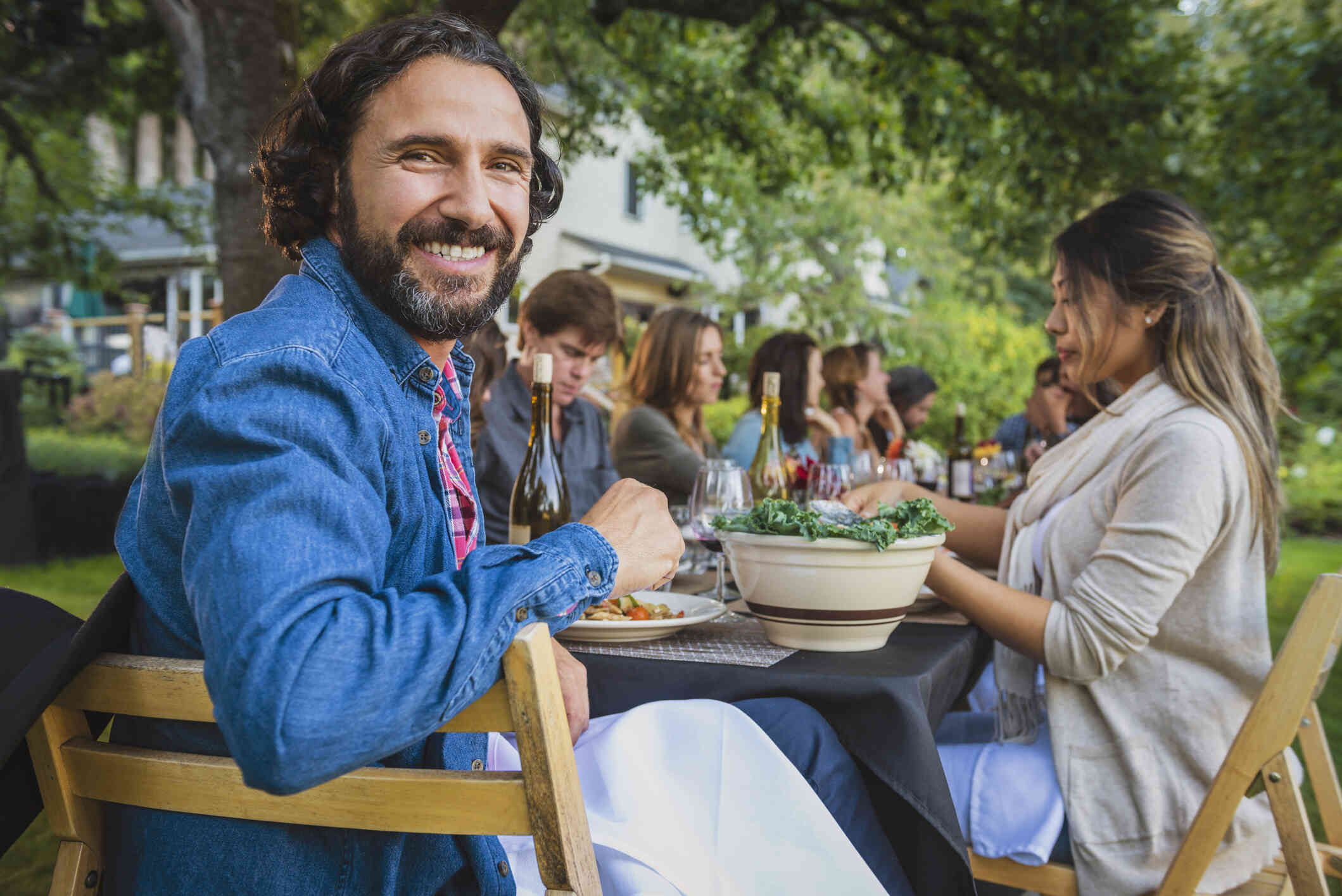 A man turns to smile at the camera while sitting at an outdoor dinner table with all of his friends.