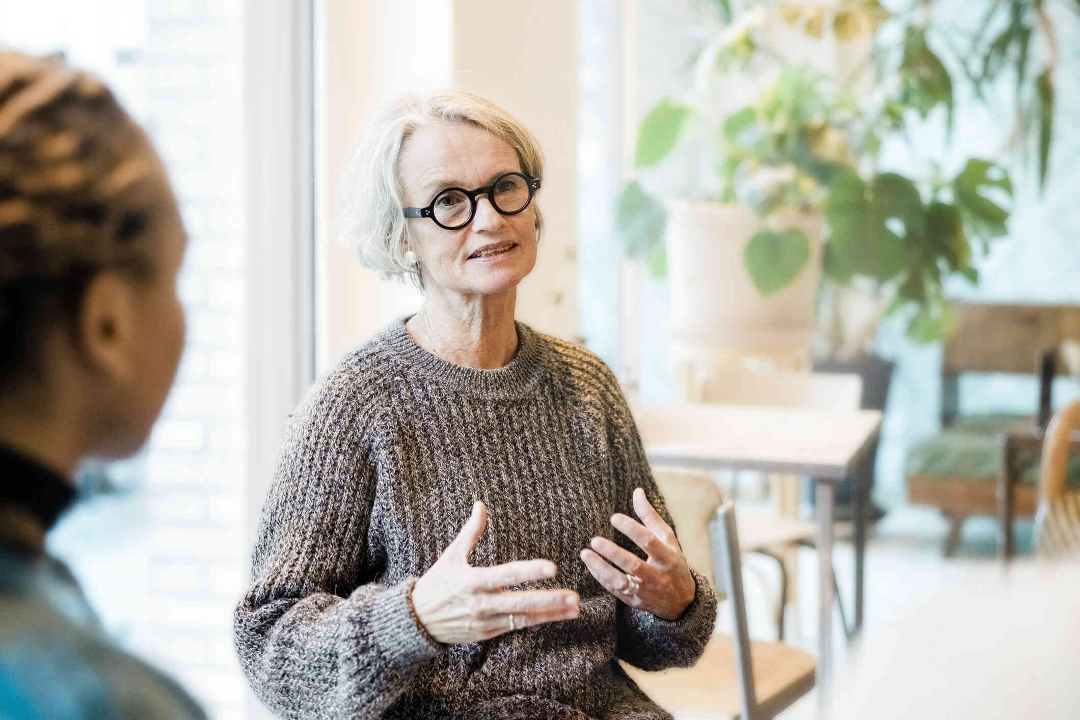 A mature woman wearing a gray sweater and glasses sits and speaks while gesturing with her hands.