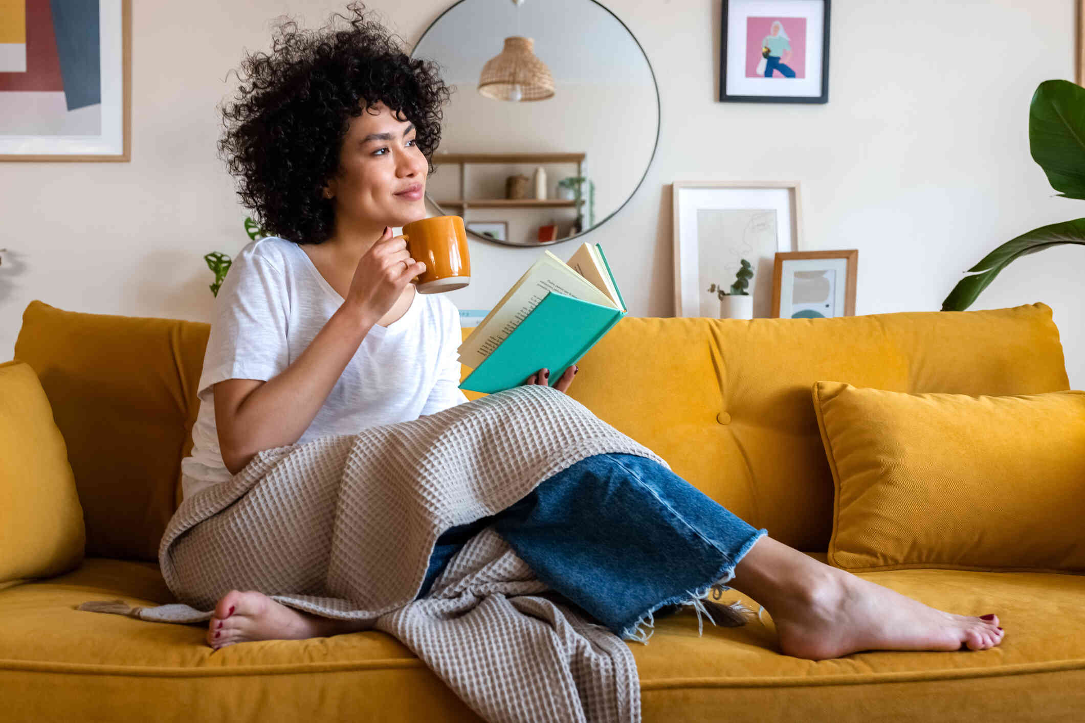A woman sits casually on her couch while with a blanket over her lap while holding a book and a cup of coffee while glancing off with a smile.