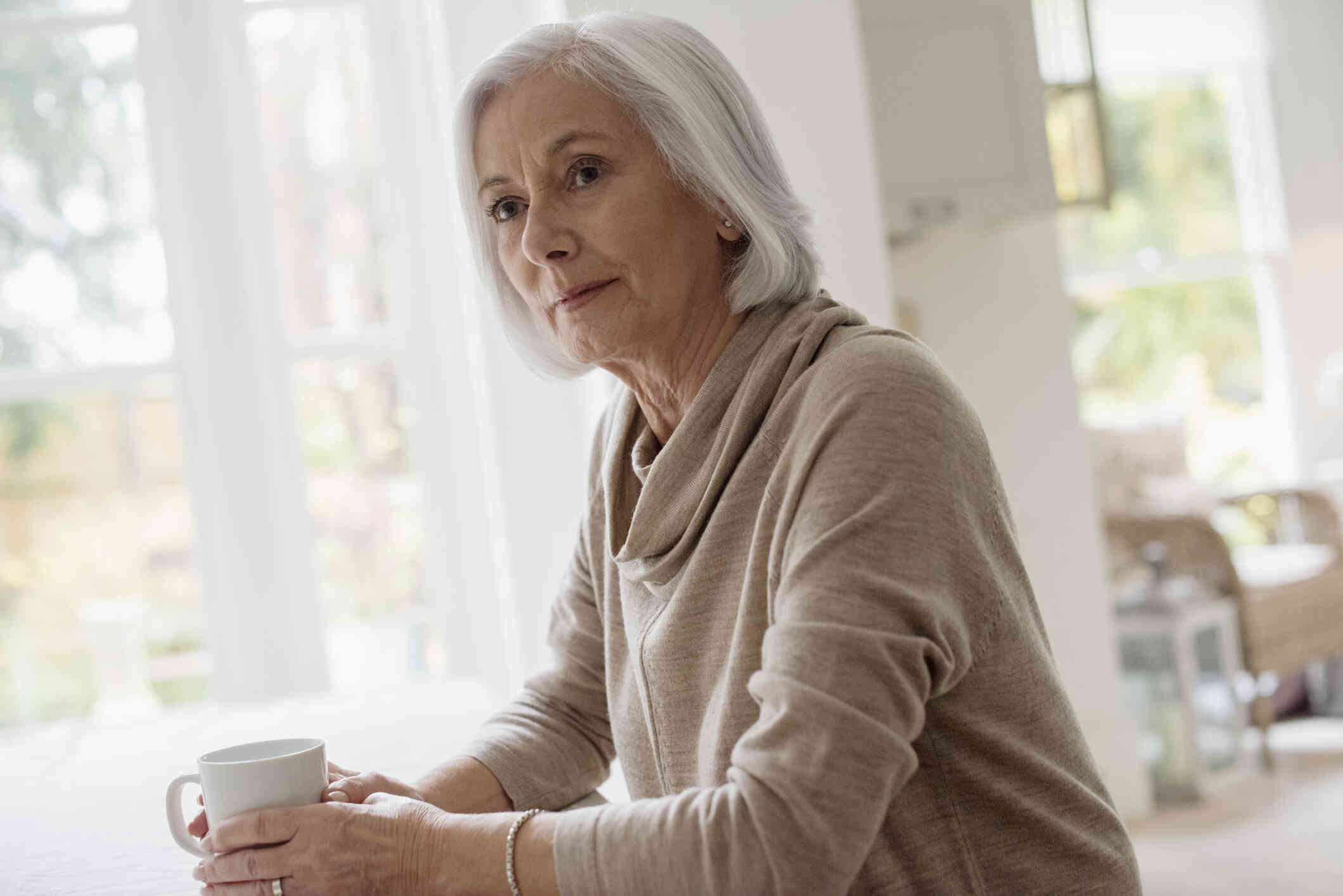 A middle aged woman in a tan sweater sits in her home and holds a white coffee mug while gazing off with a serious expression.