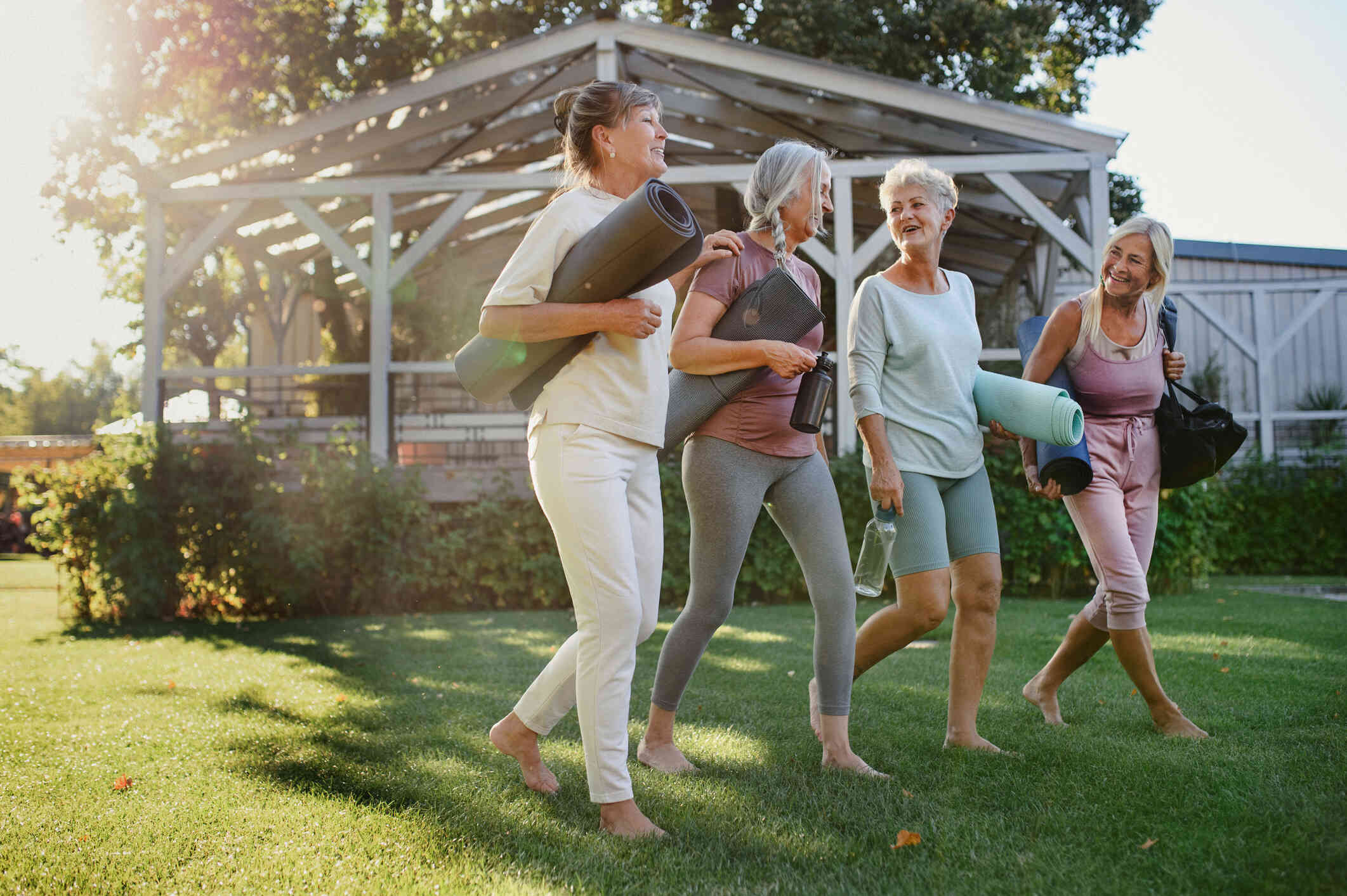 A group of adult woman walk along the grass together while holding yoga mats and chatting.