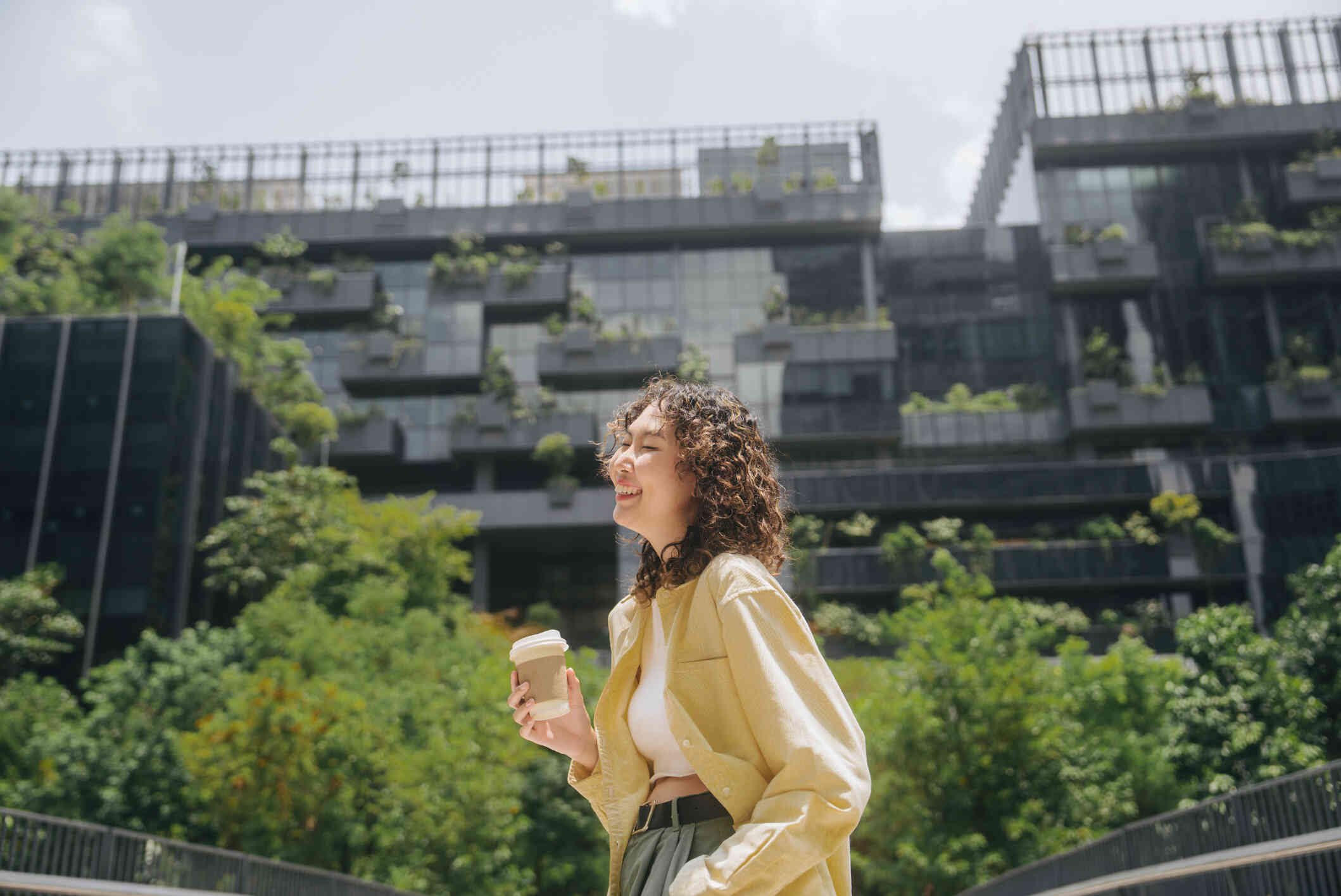 A woman stands outside on a sunny spring day while holding a to-go coffee cup while smiling brightly.