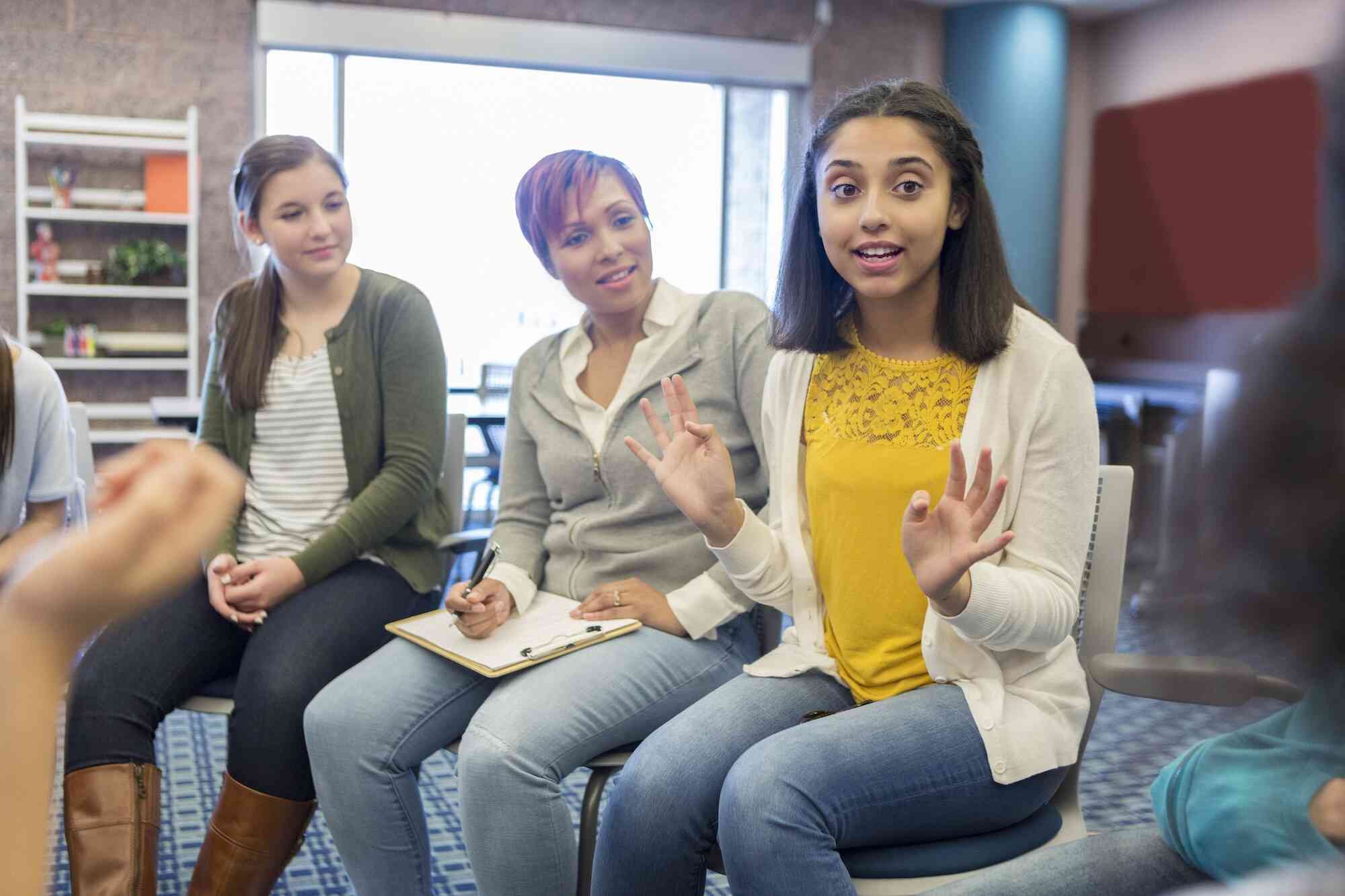 A girl in a yellow shirt sits in a chair with a positive expression while talking with a group of people