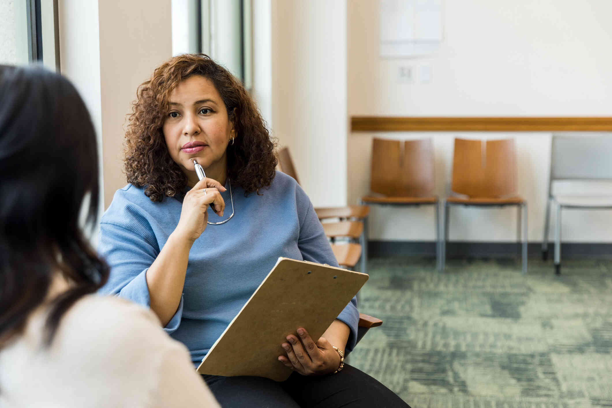 A female therapist holding a clipboard and a pen sits infront of her female patient while listening intently.