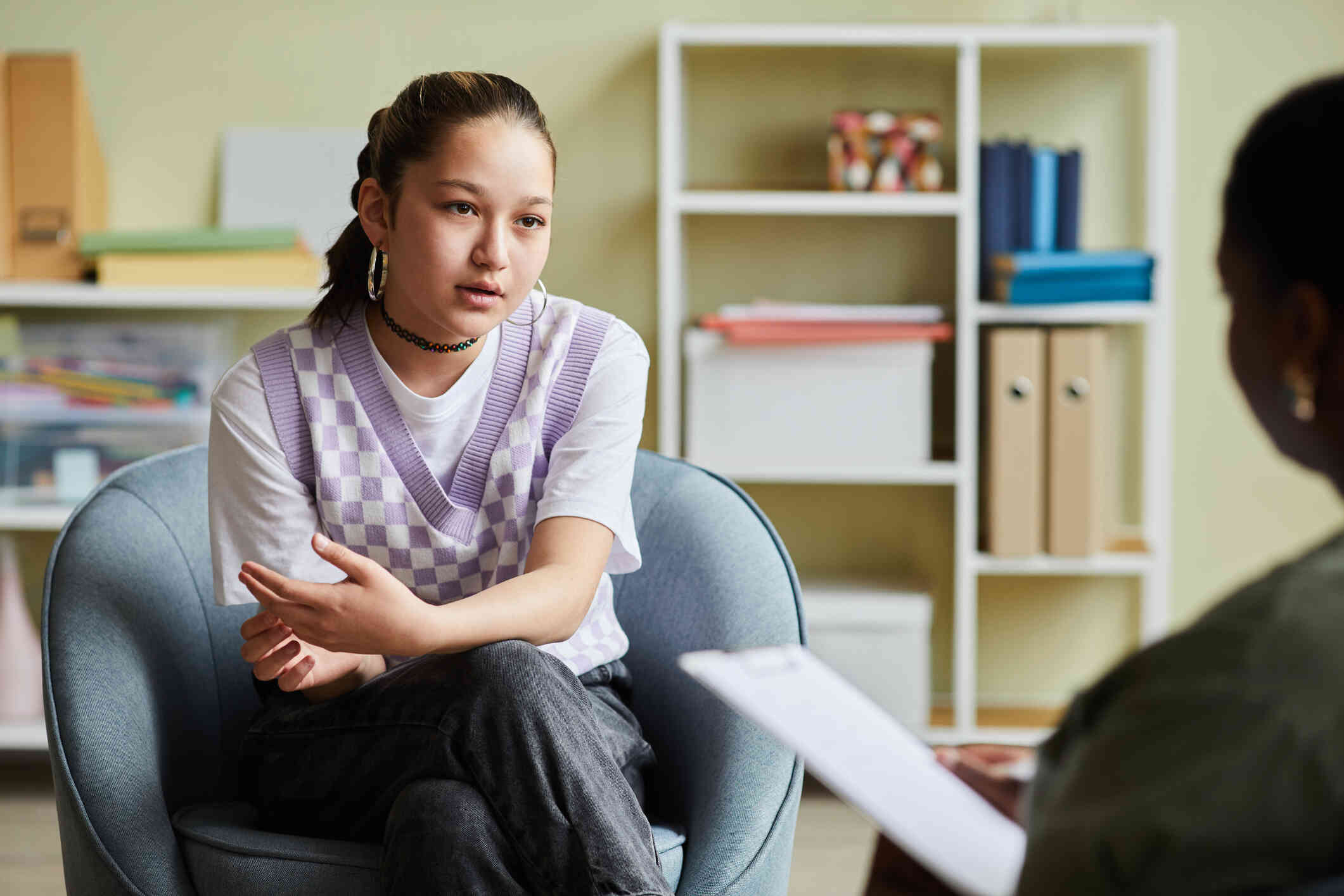 A young woman in a purple sweater vest sits in a comfy chair in an office looking at her therapist while having a discussion. The therapist sits across from her with a clipboard.