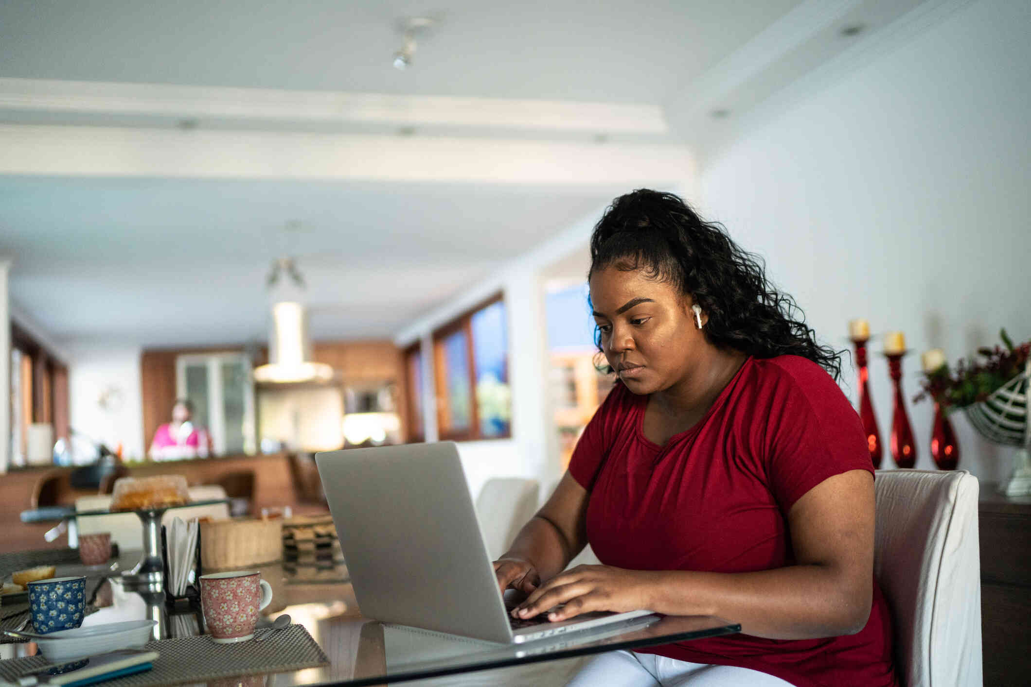 A woman in a red shirt with earpods sits at the kitchen table and types on the laptop open infront of her with a serious expression.