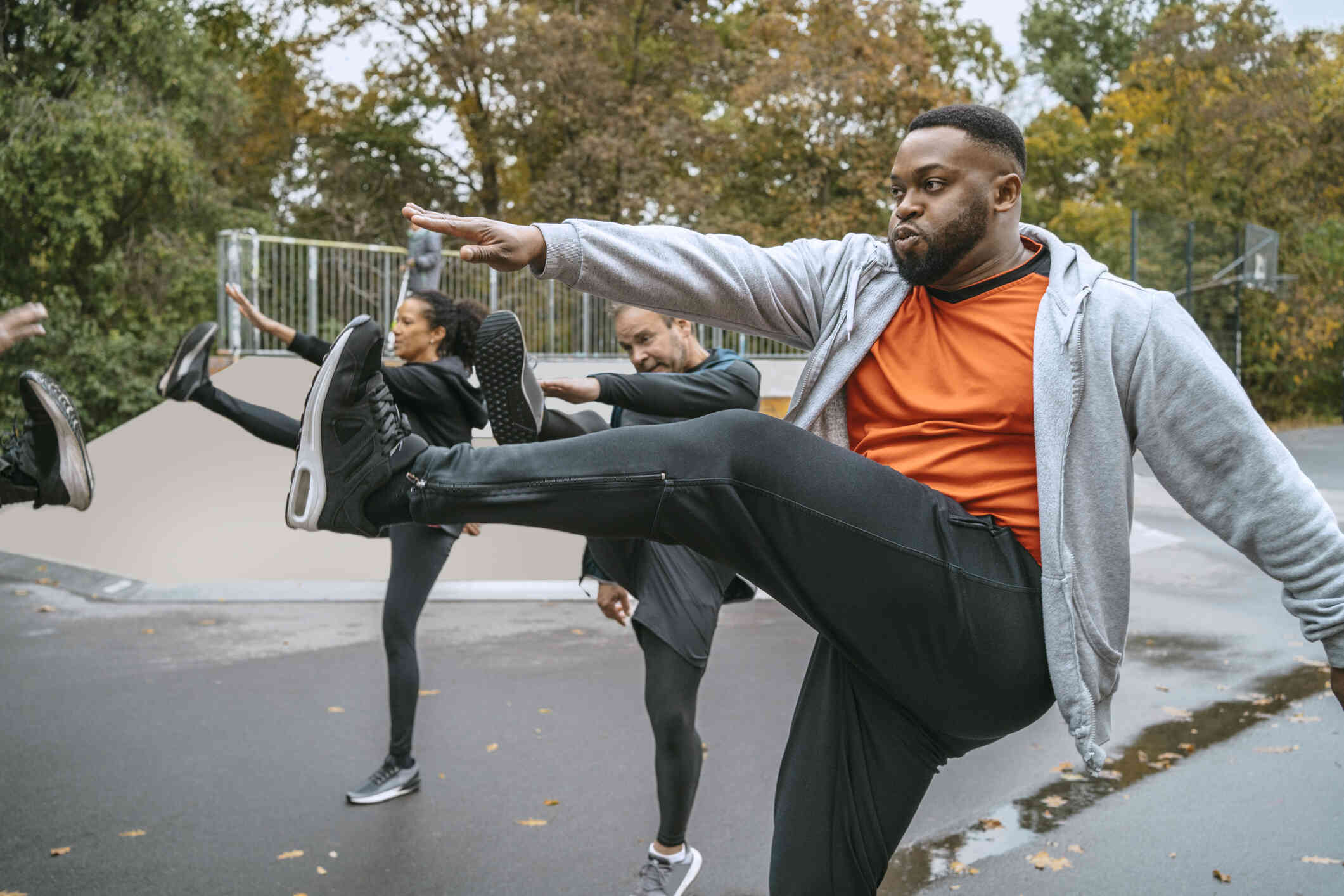 A man in winter workout attire breathes in as he stands outside on one leg and reaches a hand out towards the other leg that is extended in the air. A group of people around him do the same move.