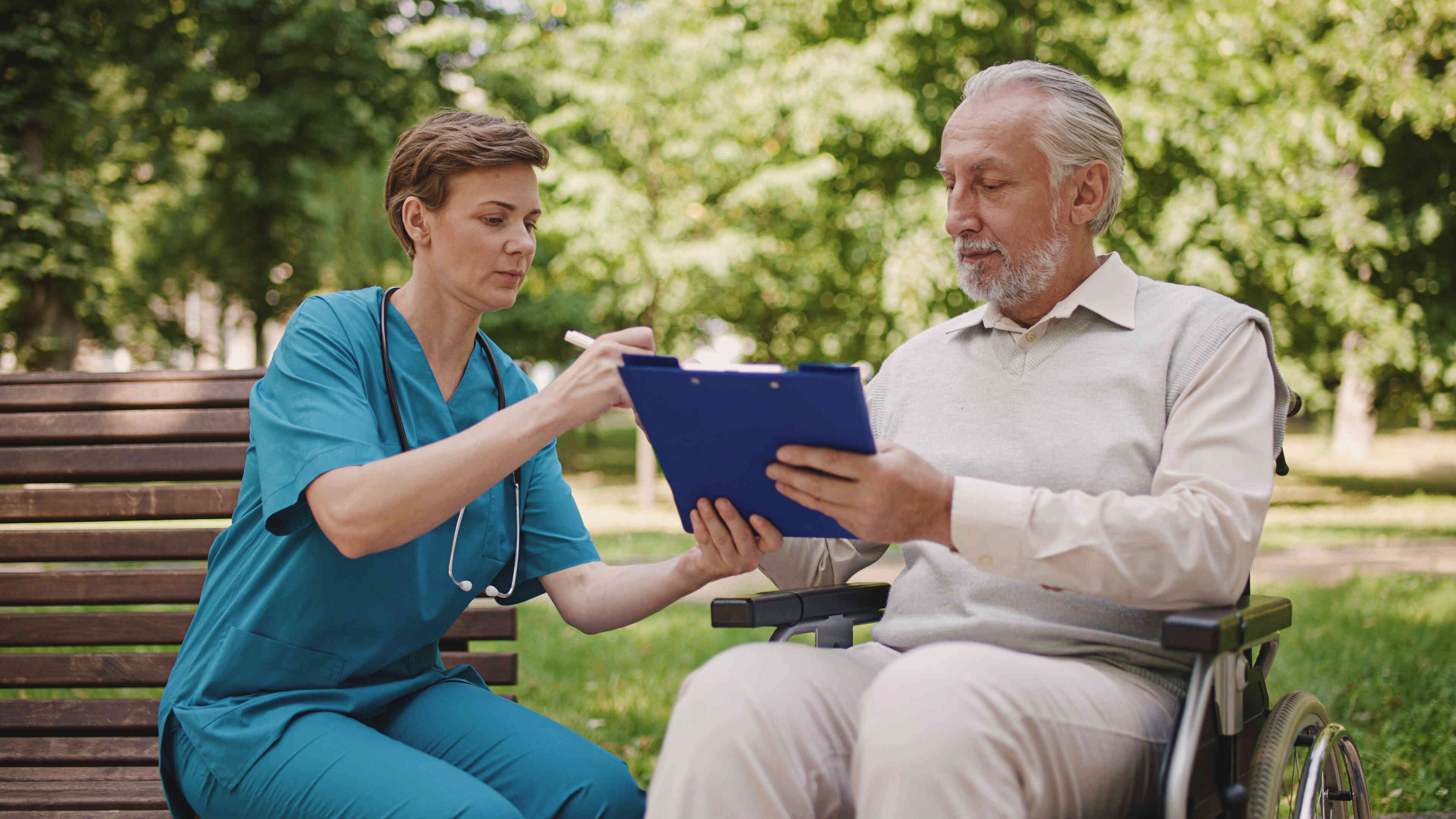 An elderly man sits in a wheelchair outside on a sunny day as a female nurse sits next to him and shows him some information on a clipboard.