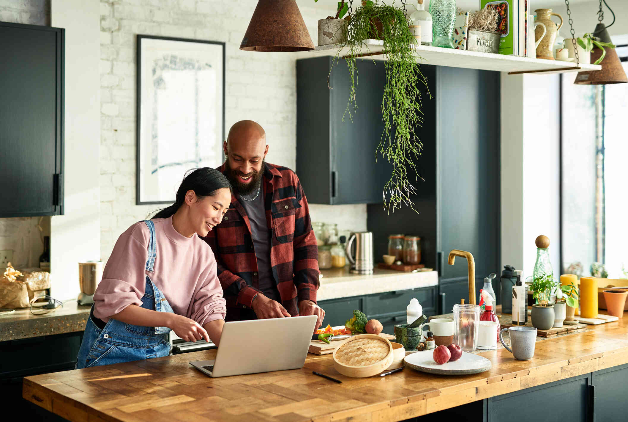 A male and female  couple stand in the kitchen and cook together while looking at the recipe on the laptop infront of them with a smile.