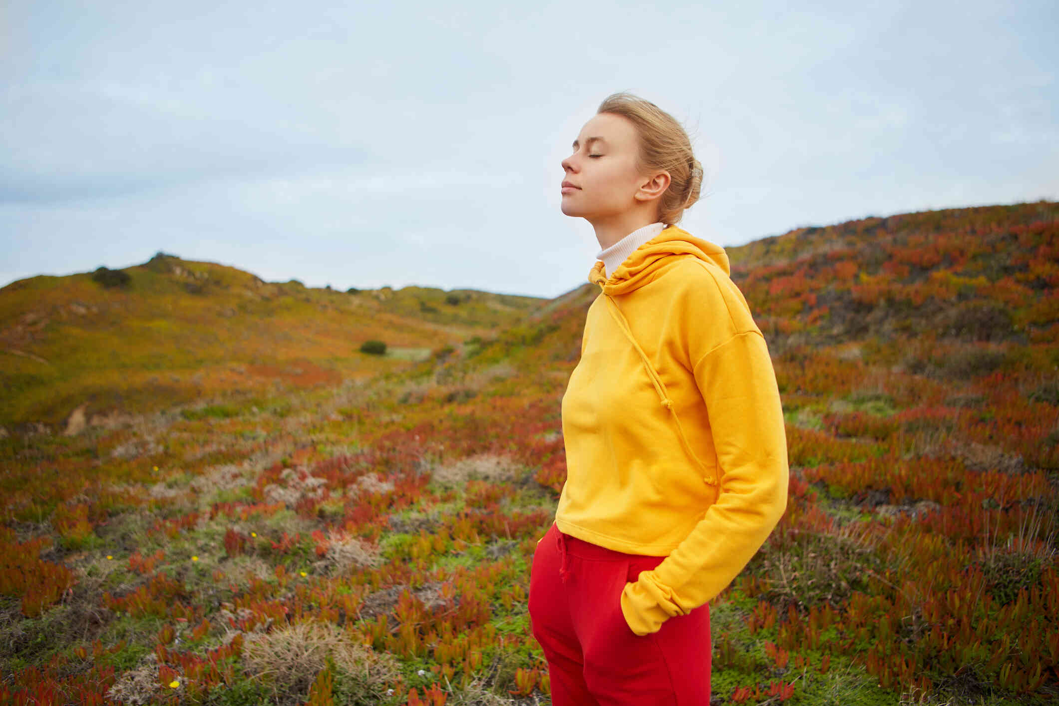 A woman in a yellow sweater stands outside on a windy day with her eyes closed.