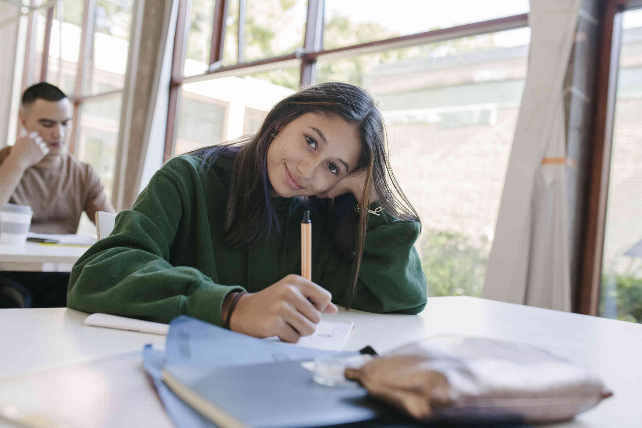 A teen girl in a green sweater sits at a table at school and writes on some papers while smiling softly at the camera.