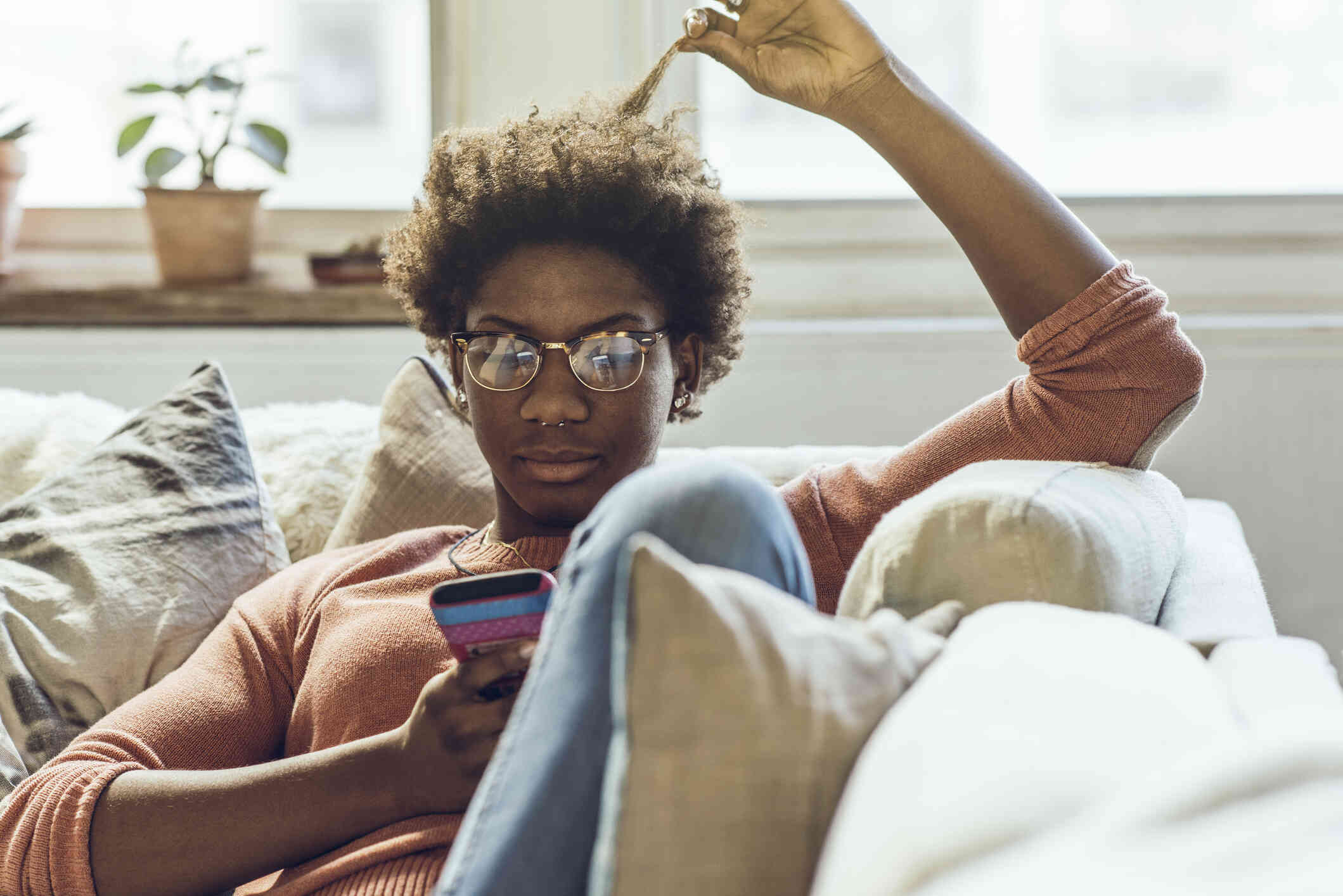 A woman with glasses sits casually on her couch and plays with her hair while looking at the cellphone in her hand.