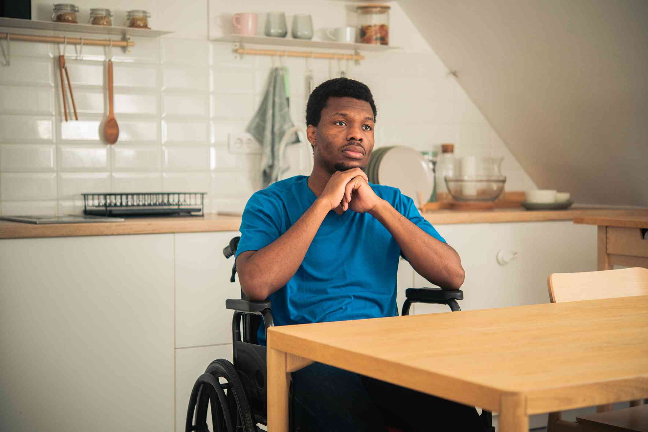 A man in a blue shirt sits in a wheelchair at a wooden table in a kitchen and glances off with a sad expression.