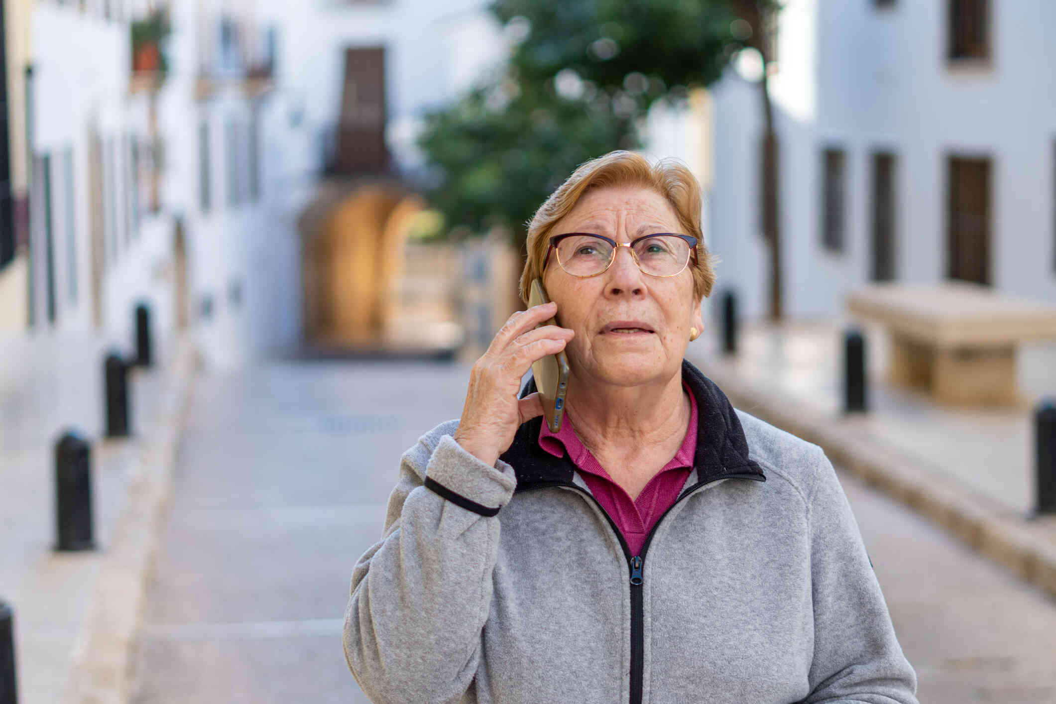 A mature woman in a grey hoodie walks outside on a coudy day and talks on the phone while staring off with a worried expression.