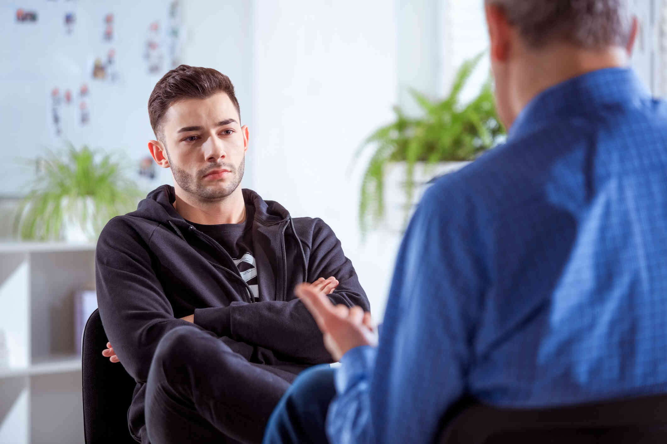 A man in a black jacket crosses his arms while sitting in chair across from his therapist during a therapy session.