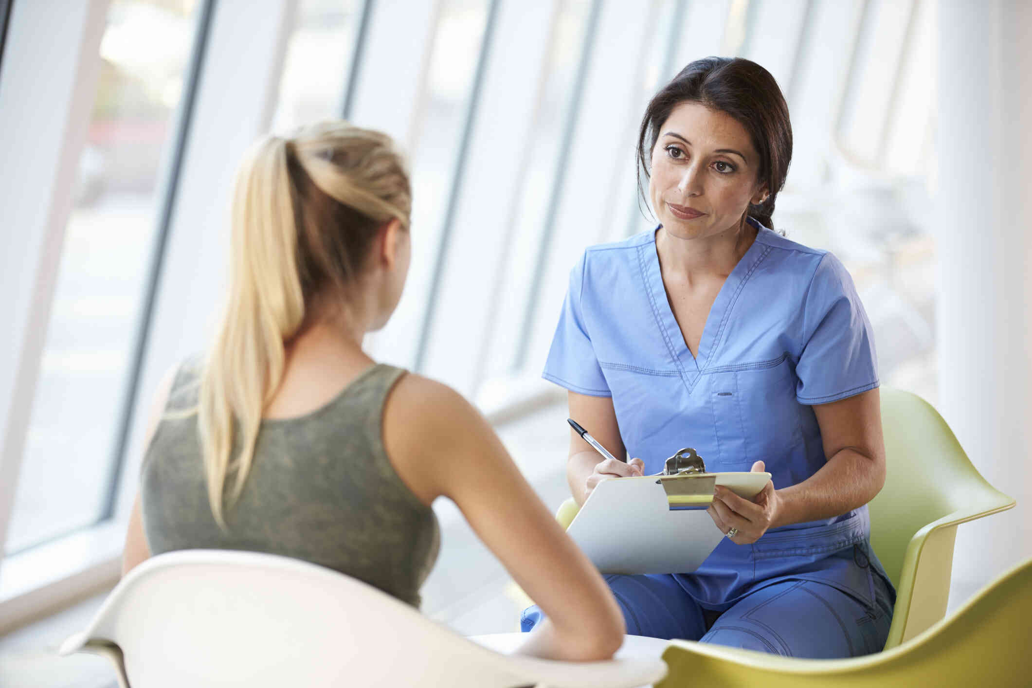 A young clinician with a neutral expression in blue scrubs talks to a young girl with a green shirt while taking notes