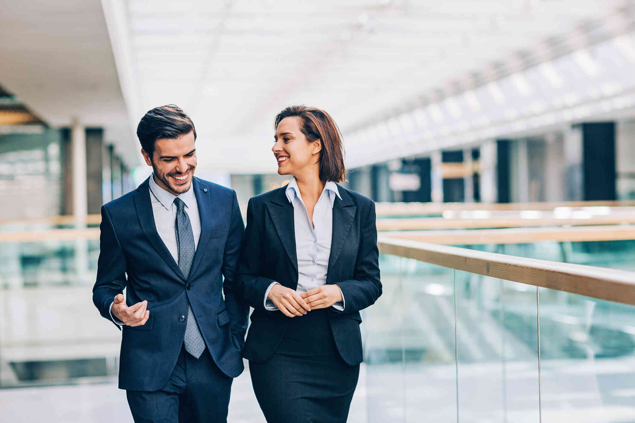 A male and female coworker in buisness attire walk side by side down a hallways while chatting and smiling.