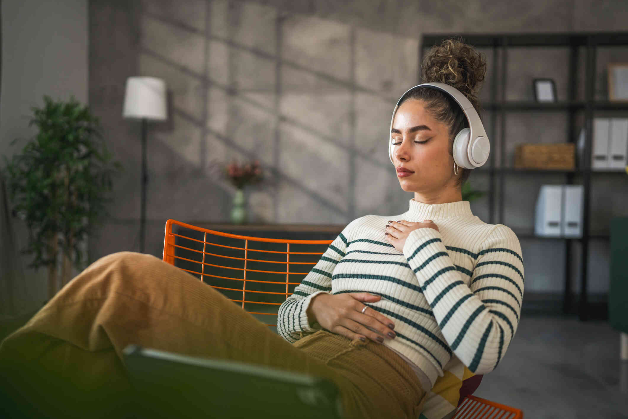 A woman sits in a chair with her eyes closed while wearing headphones and places one hand over her heart and one over her stomach.