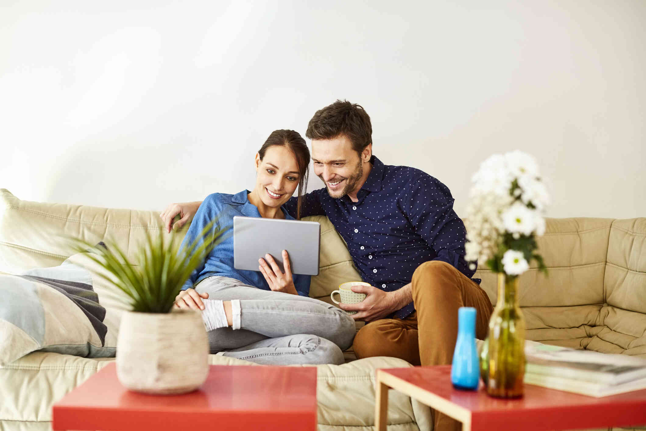 A male and female couple sit sie by side casually on their couch as they both smile and look at the tablet in the womans  hand.
