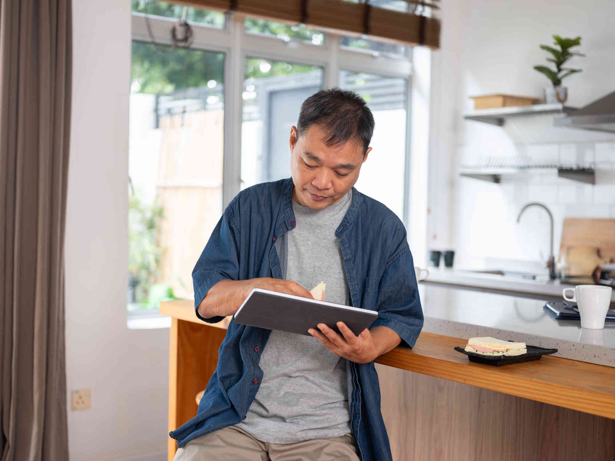 a middle aged man in a blue shirt stands in his kitchen and taps on the tablet in his hand with a serious expression.