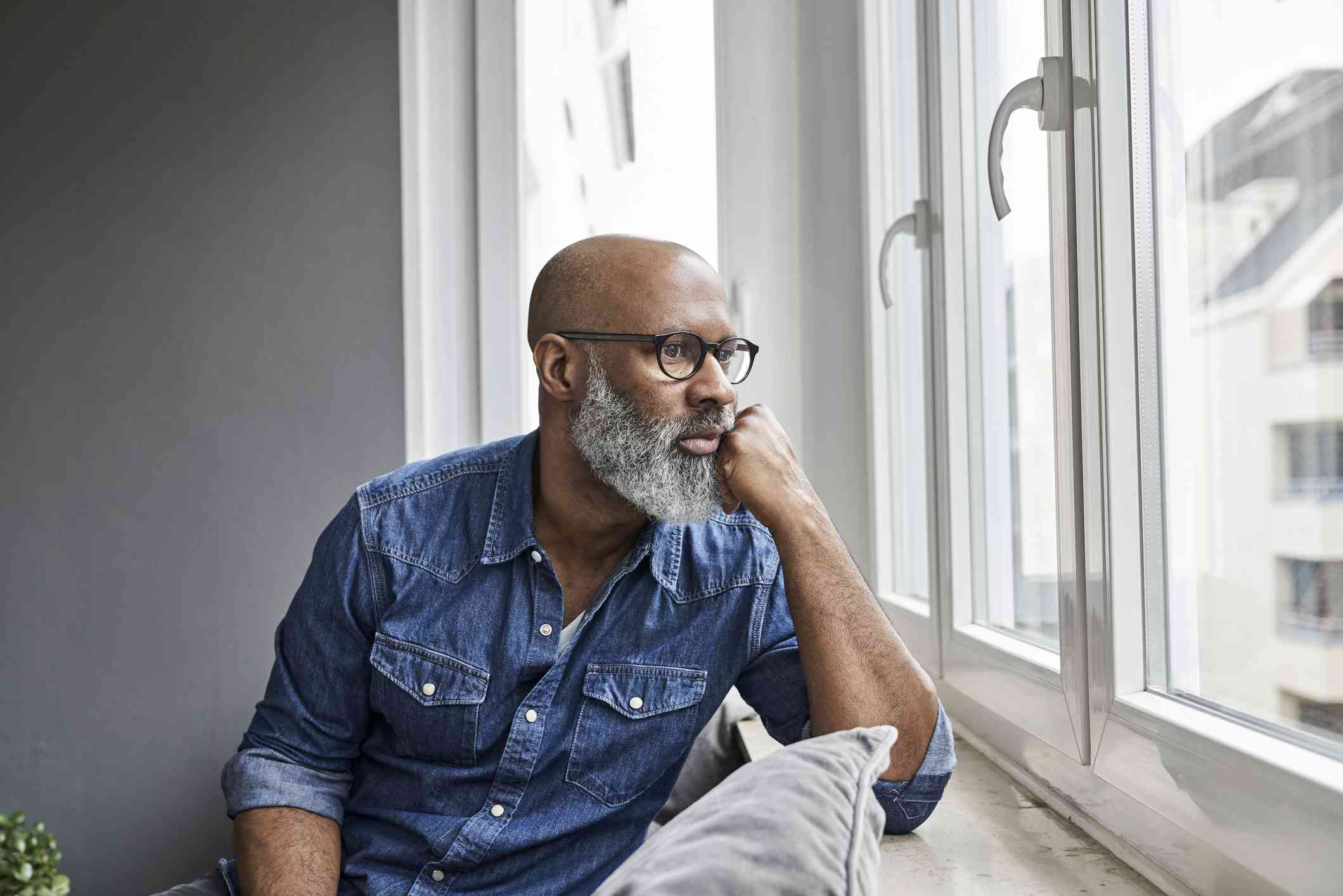 A middle aged man in a blue jean button down shirt sits on the couch near a window and gazes out whiel deep in thought.