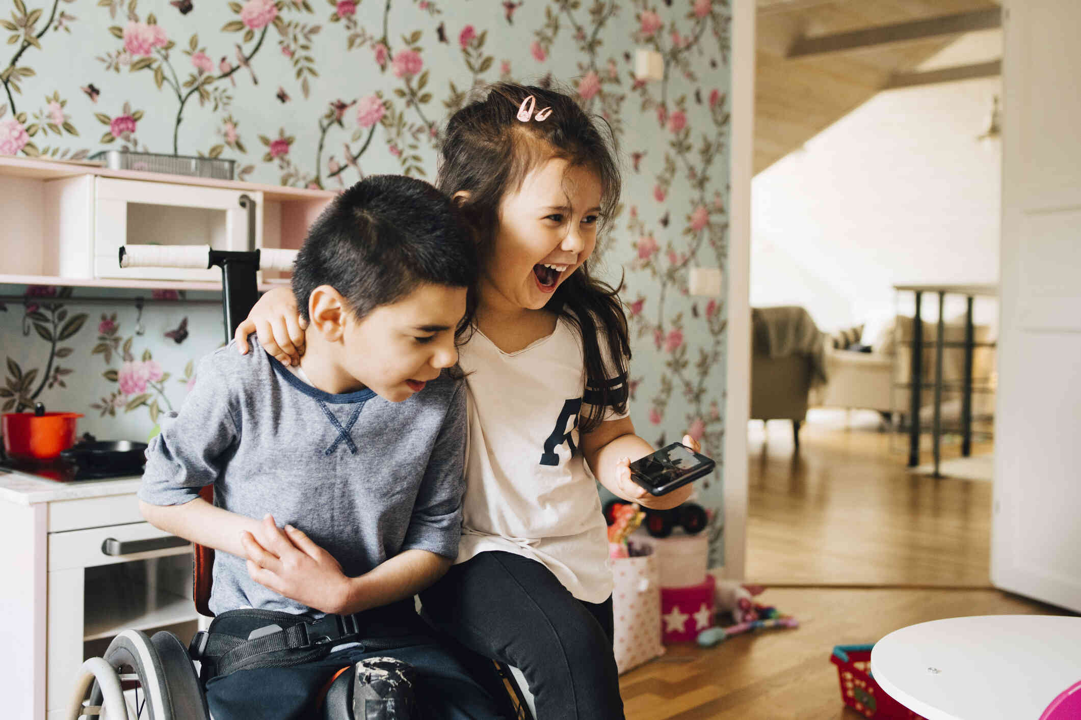 A young boy and a young girl smile as they stand next to each other in a children's room and look at a phone the girl is holding in her hand.