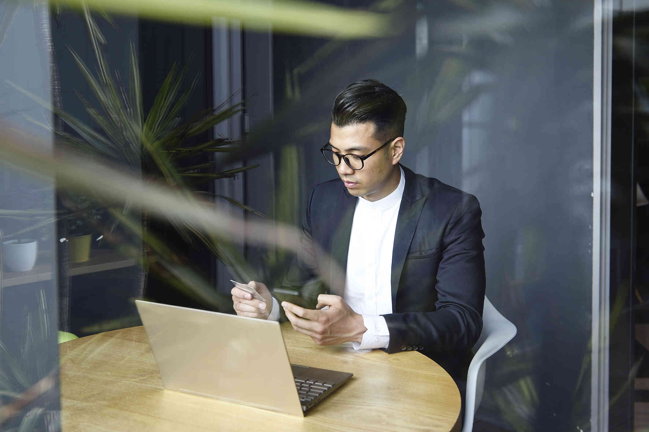 A man in a blazer sits at a table at work wth  his laptop open infront of him while he holds a cellphone in one hand and a buisness card in the other.