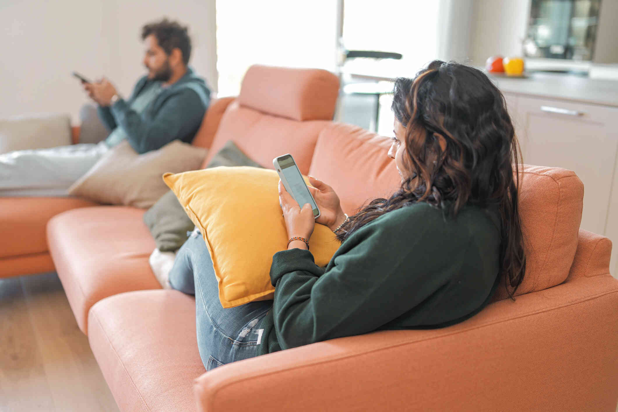 A girl with long dark hair sits on one end of the couch looking at her phone with a pillow on her lap while a man in the background sits at the other end of the couch on his phone.