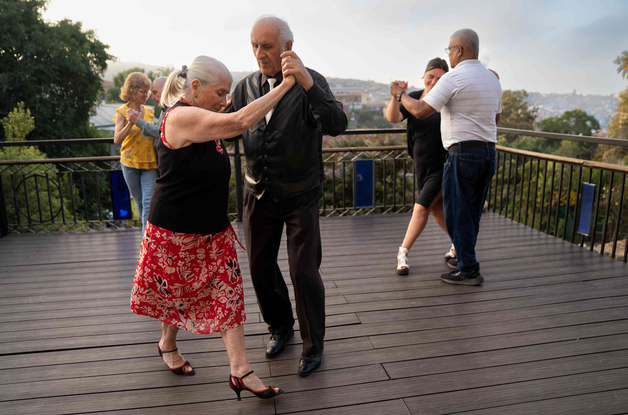 A mature woman wearing a long dress dances outside on a deck with a mature man wearing leather jacket. Two other couples dance together in the background.