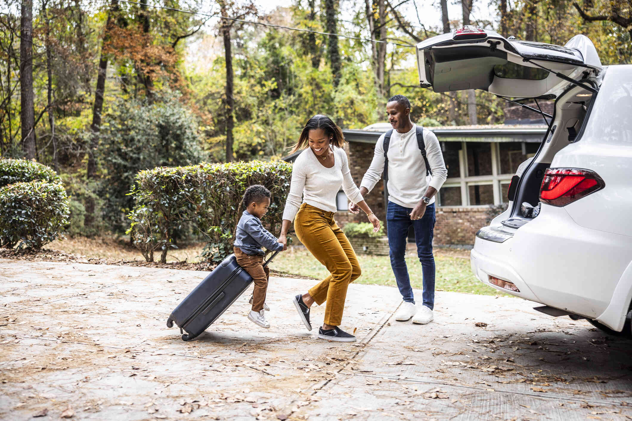 A young boy smiles as he rides on a rolling suitcase as a woman pulls it towards a car with it's trunk open. A man stands outside by the trunk ready to grab the suitcase.