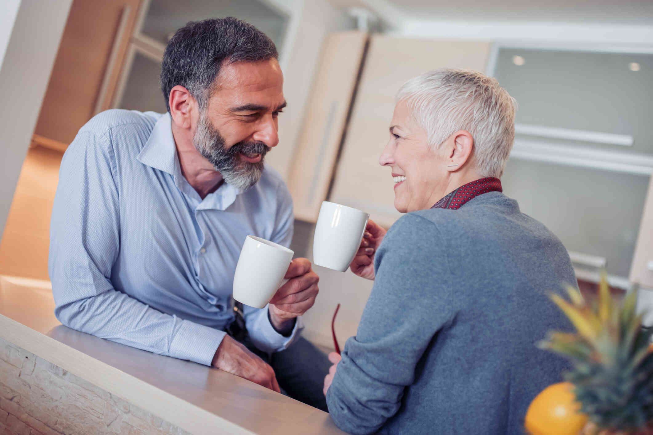 A middle aged male and female couple smile at one another while sitting acroos from each other at a table and holding cups of cofee.