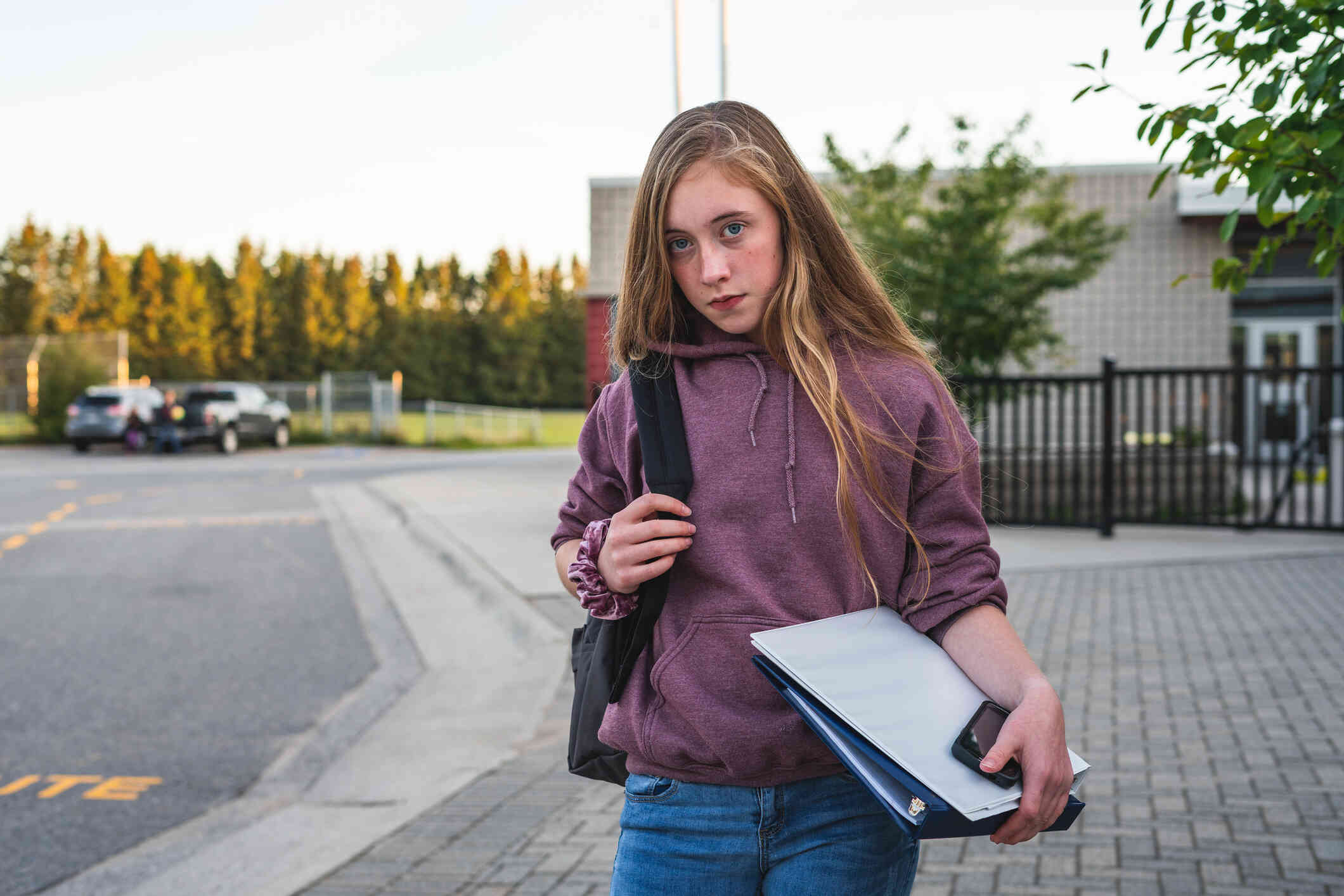A teenage girl in a purple hoodie stands outside of her school with her backpack and looks at the camera with a serious expression.