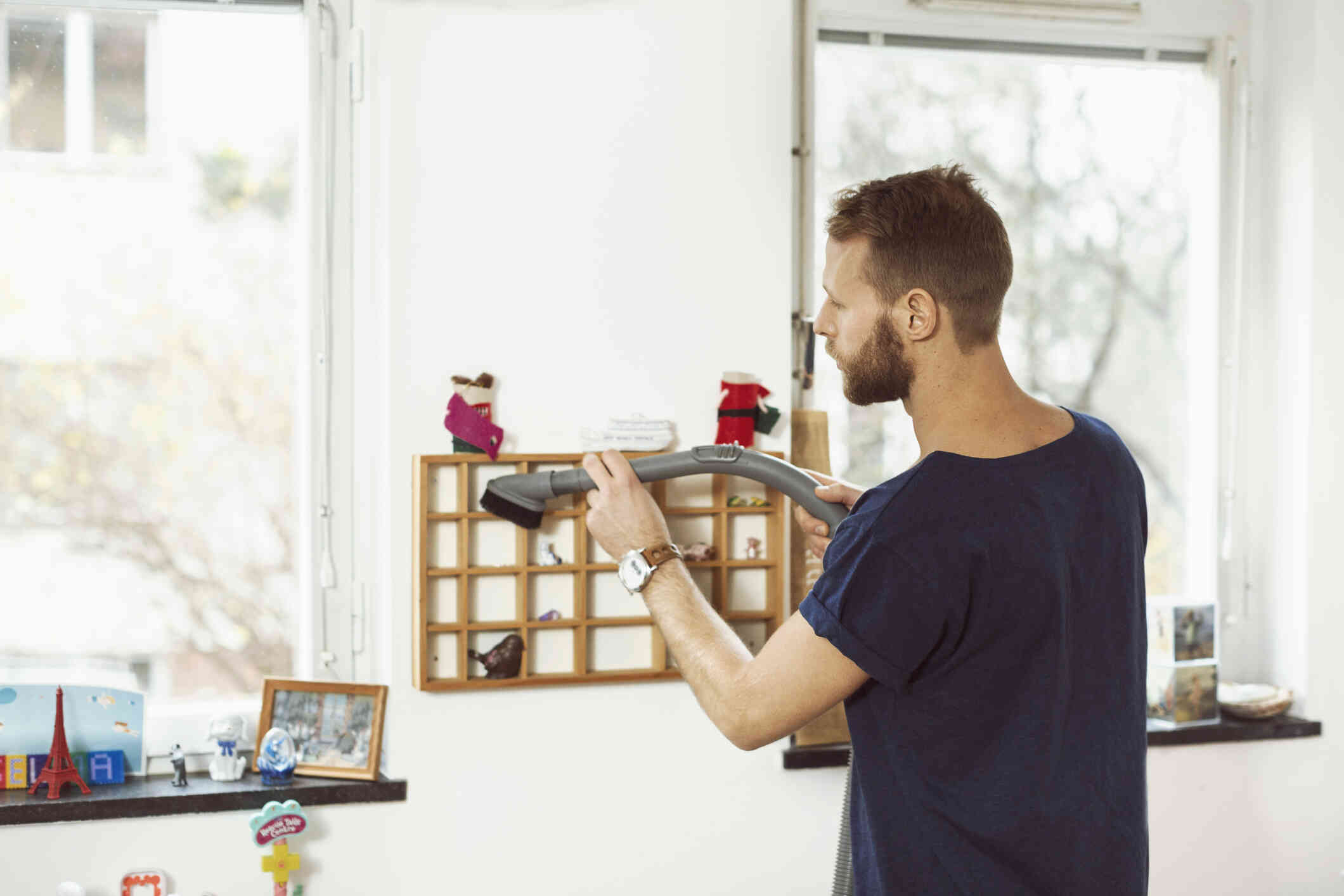 A man in a blue shirt vacuums a wooden shelf in a kids room.