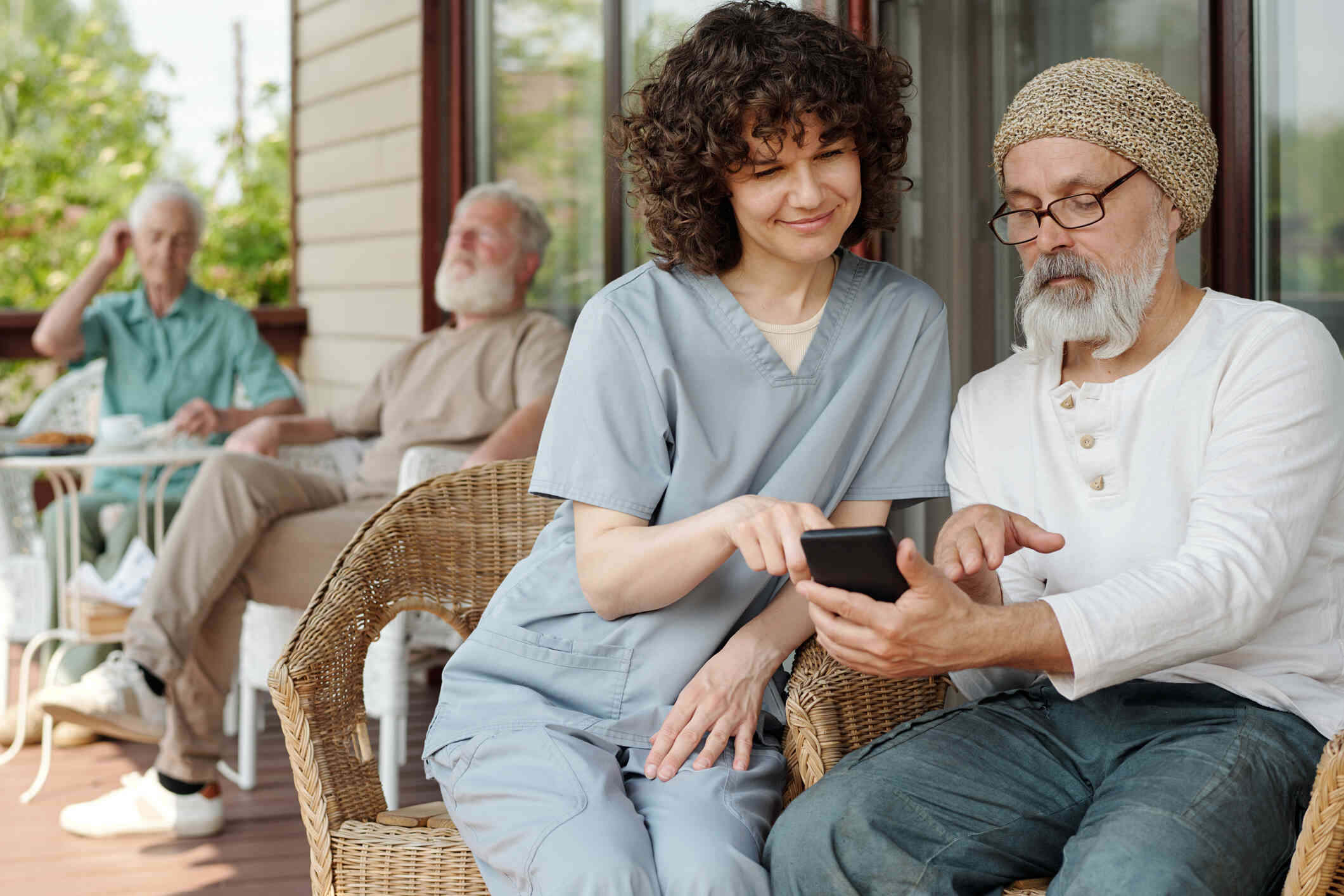 An elderly man sits on a porch in a whicker chair with his cellphone as a female nurse sits next to him and points at his phone with a smile.