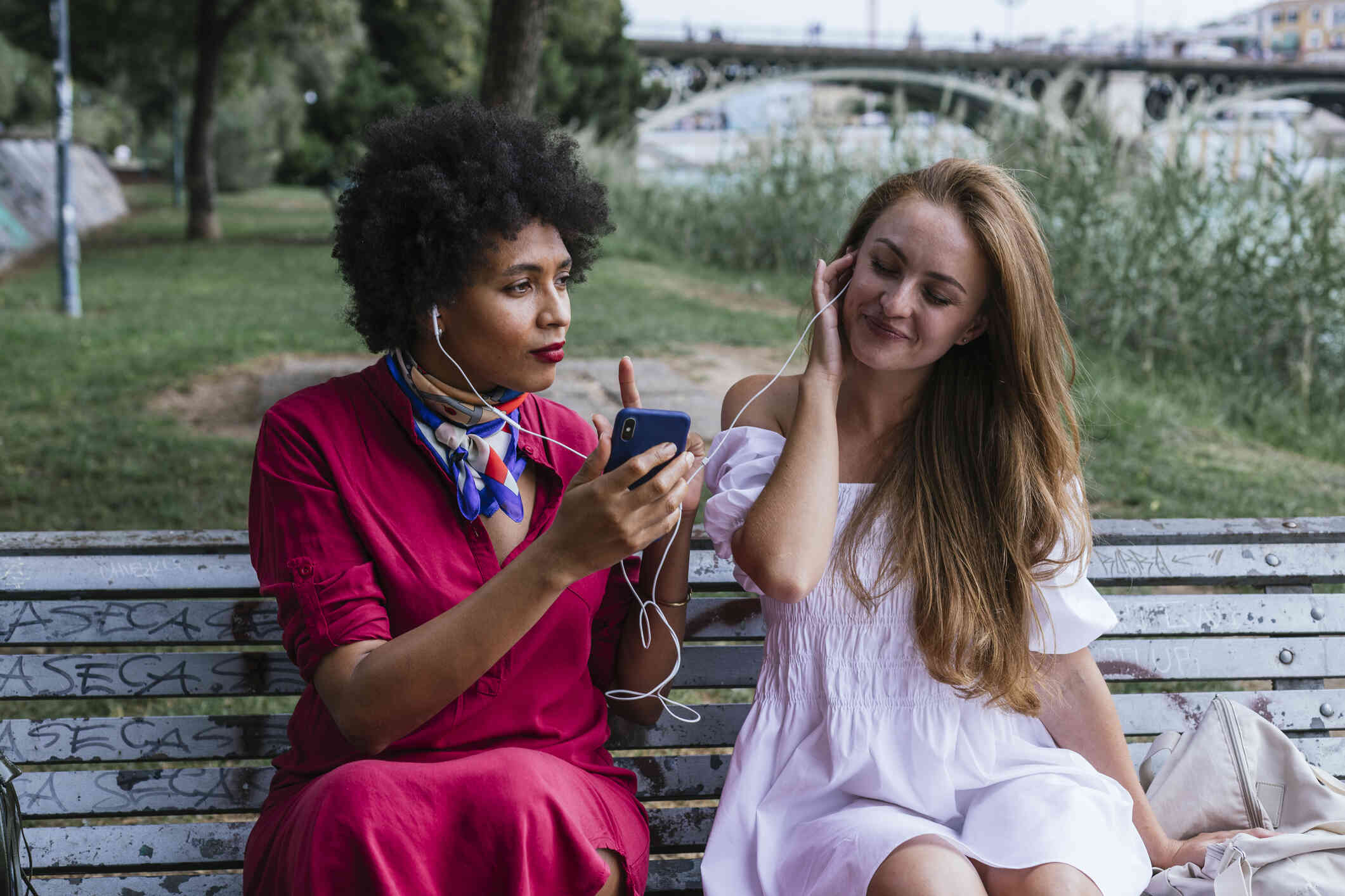 Two adult female friends sit on a park beck together while listening to music from the same headphones.