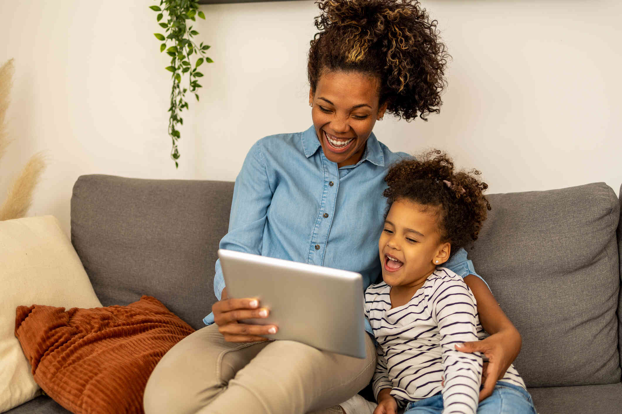 A mom sits on the couch with her young daughter as they both smile down at the tablet in the moms hand.