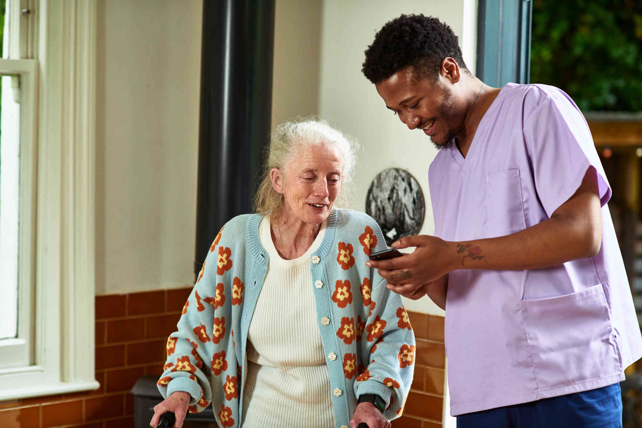 A male nurse in purple scrubs shows an elderly woman his phone as they both smile.