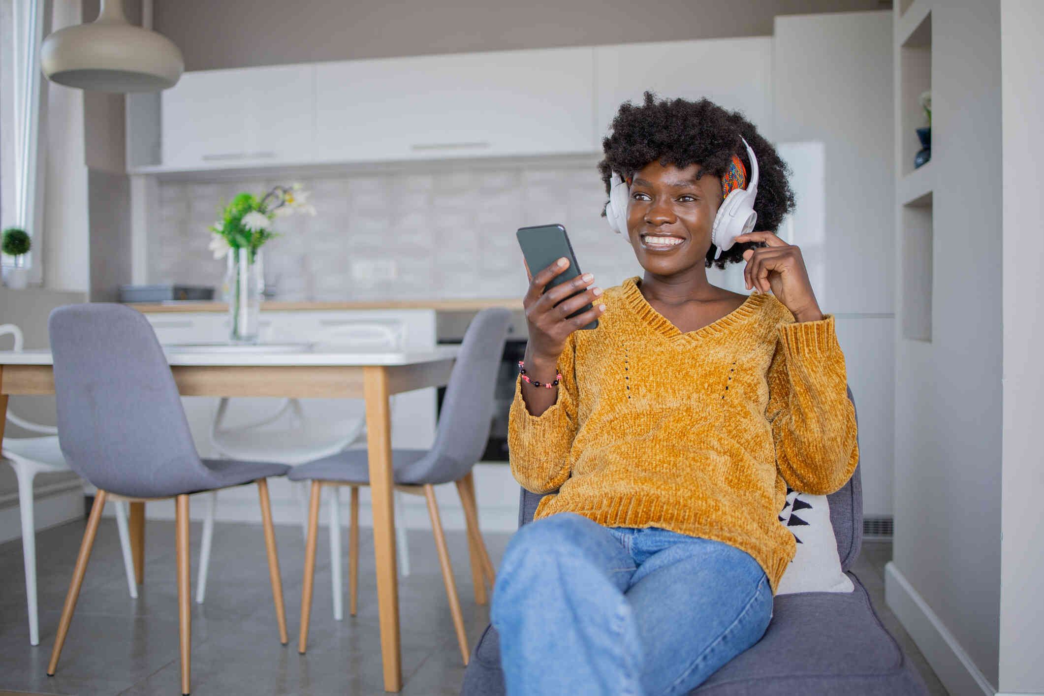 A woman wearing a yellow sweater and headphones smiles as she holds a cellphone in her hand.