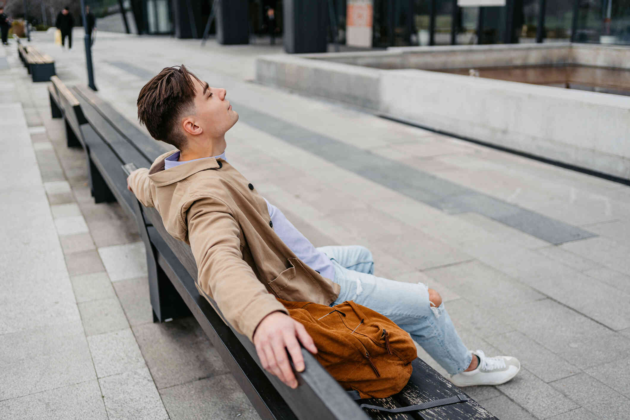 A man in a brown coat sits on a bench in the city with his arms resting on the back of the bench as he closes his eyes and lifts his head.