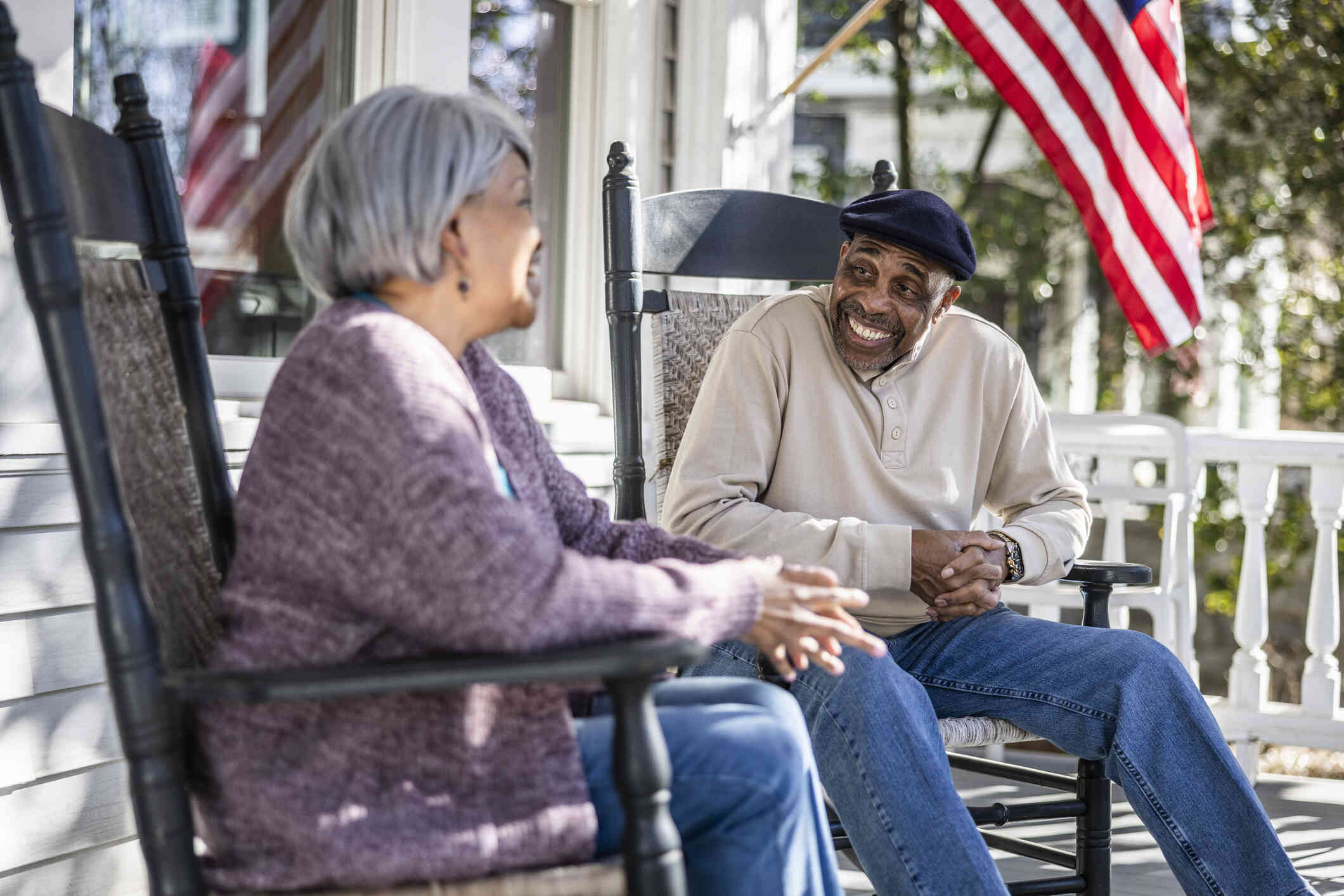 An elderly male and female couple sit in rocking chairs on the front porch and smile while chatting with one another on a sunny day.
