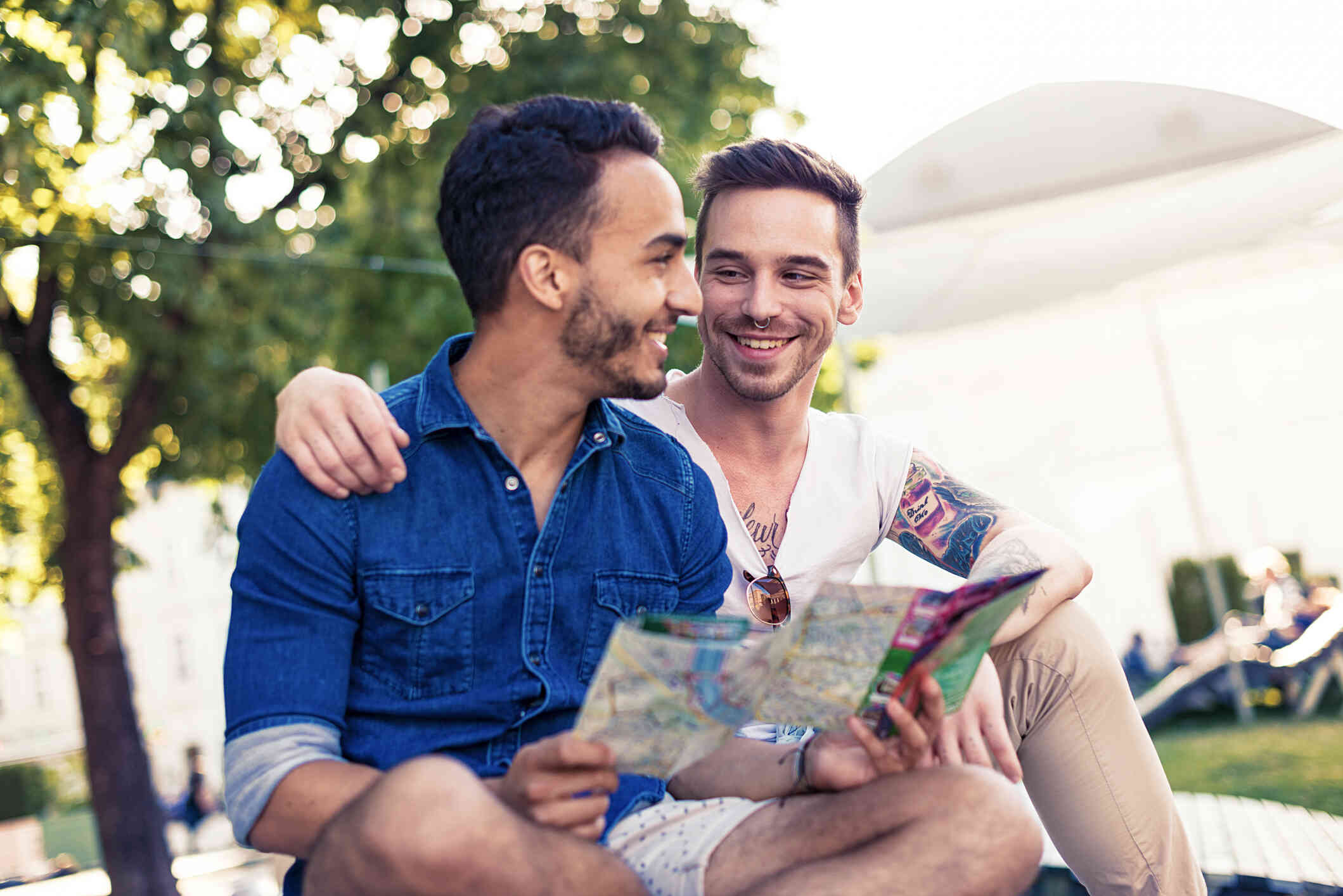 A male couple sit outside on a blanket and smile at one another while looking at a park map.