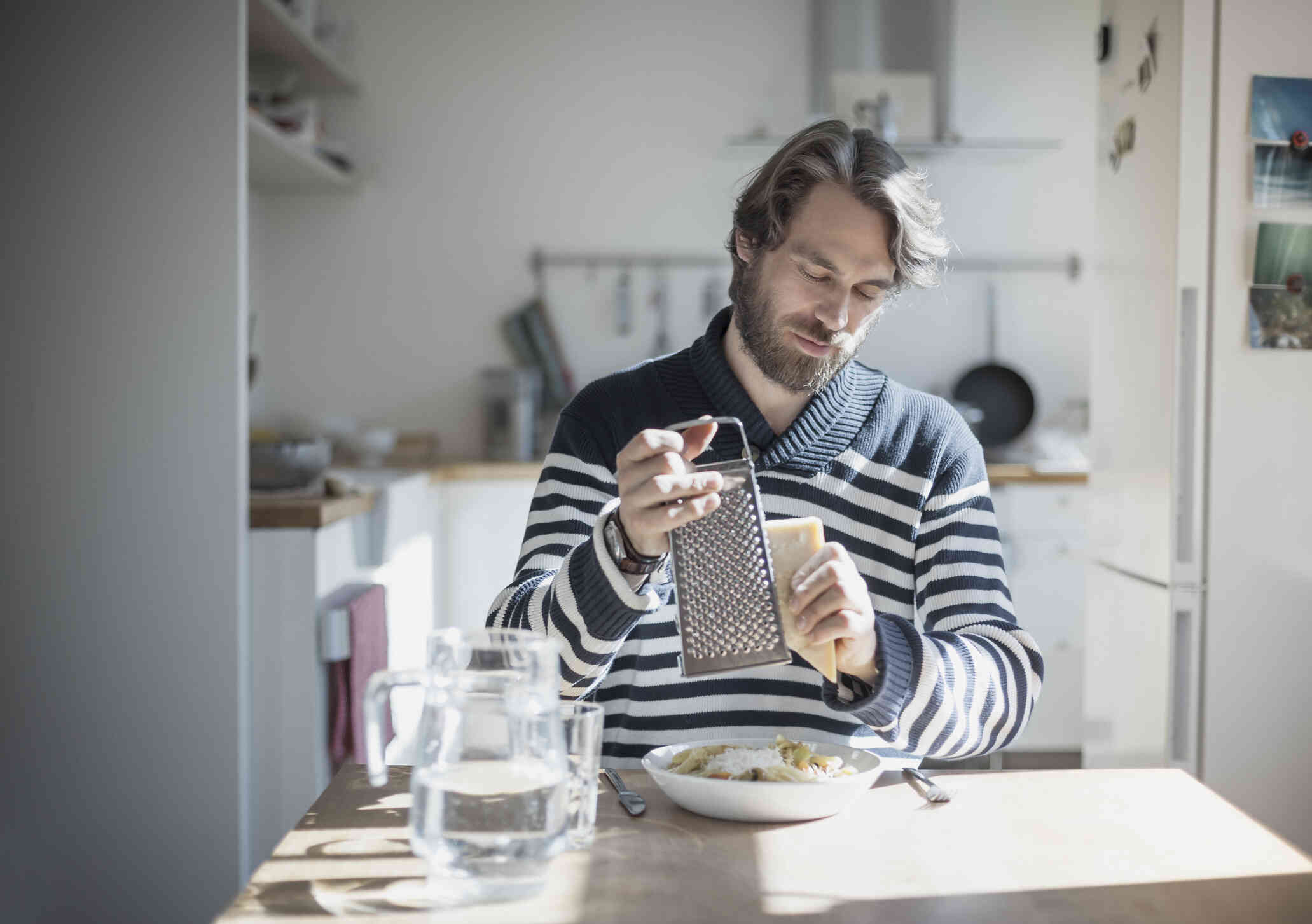 A man in a striped shirt sits at his kitchen table and grates cheese onto his food.