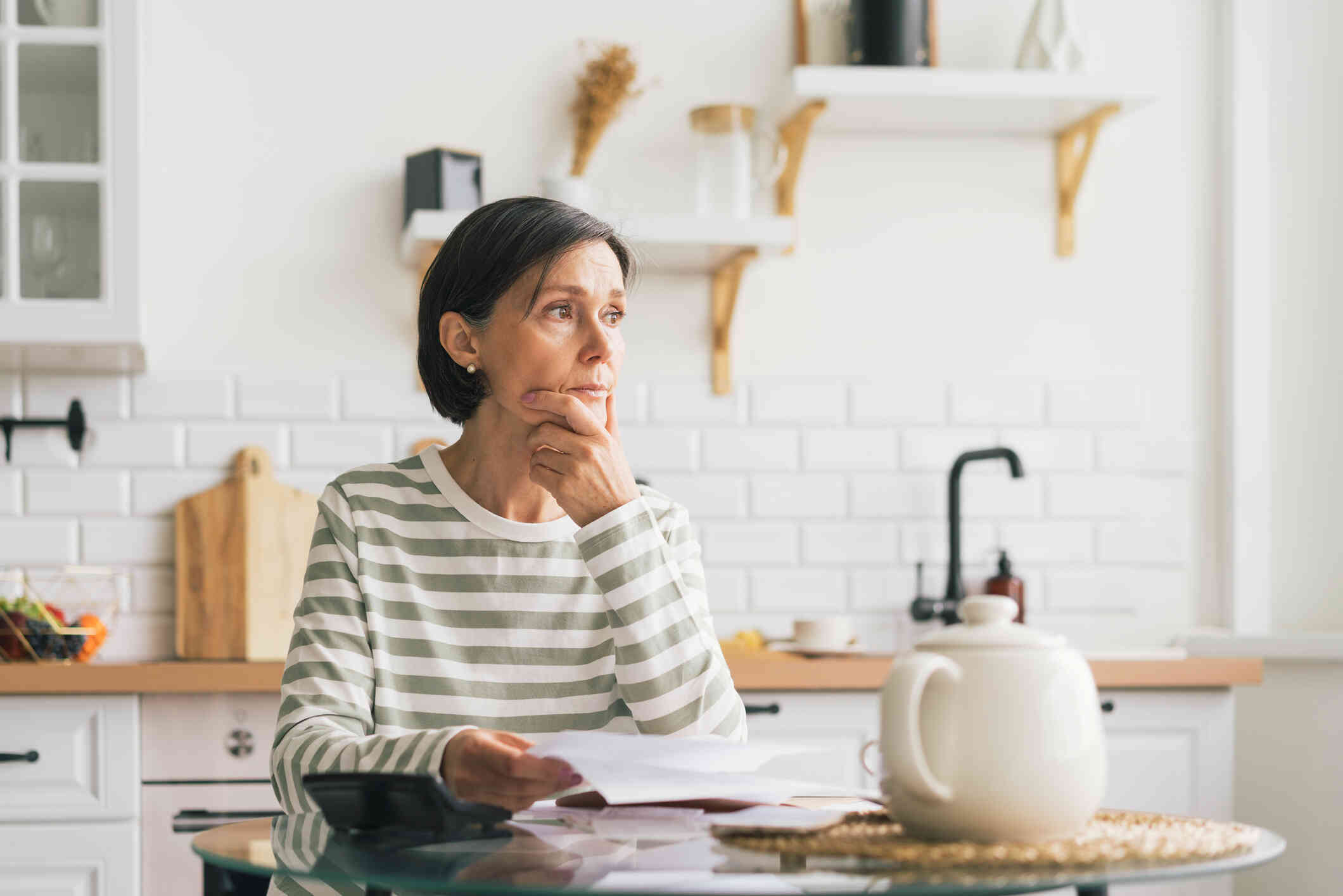 A middle aged woman sits at her kitchen table with papers in her hands as she presses her other hand to her face and gazes off sadly.