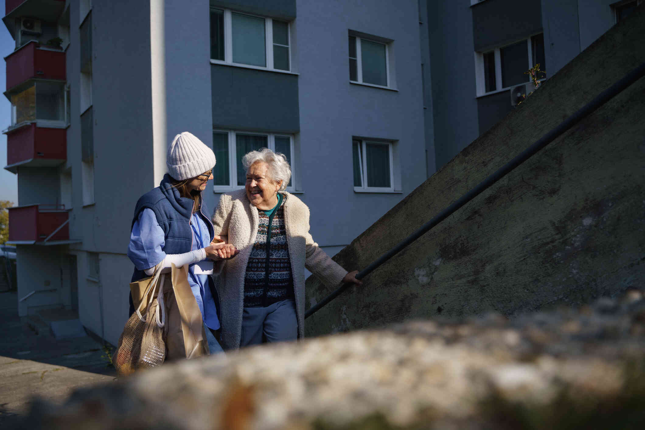 A female nurse in blue scrubs and jacket helps an elderly woman walk up some cement steps while outside on a cold day.