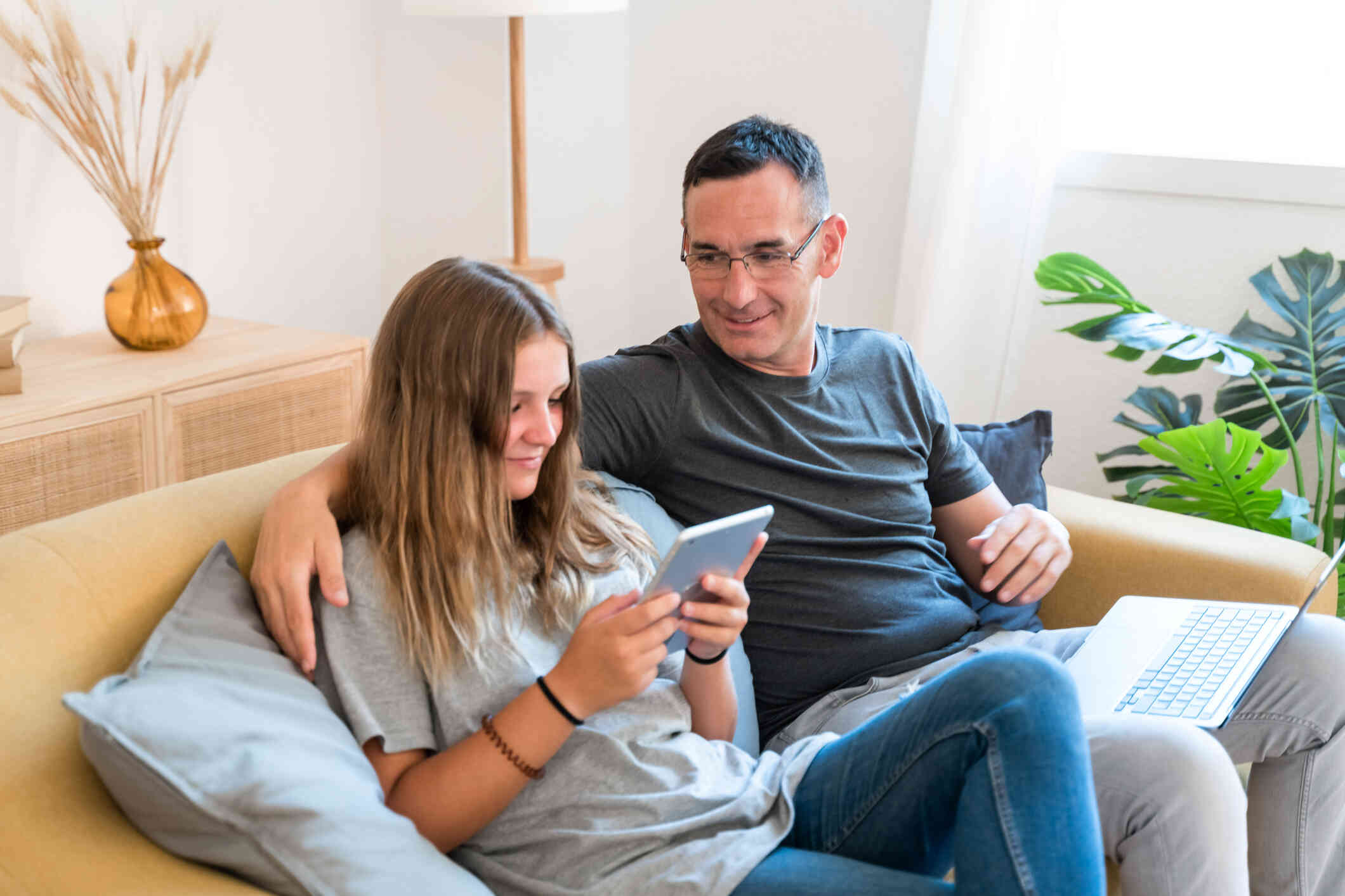 A man in a grey shirt and glasses sits on the couch with his daughter while she looks at her tablet 