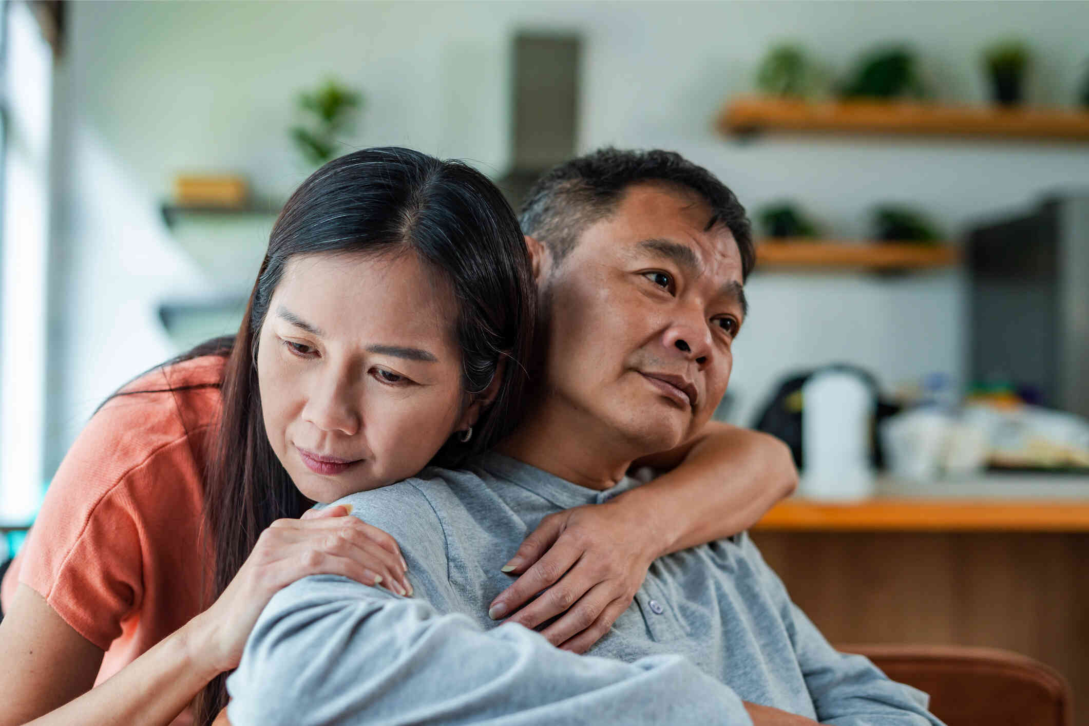 A woman sits behind a man and wraps her arm around  her male partners neck lovingly as they  both gaze off with sad expressions.