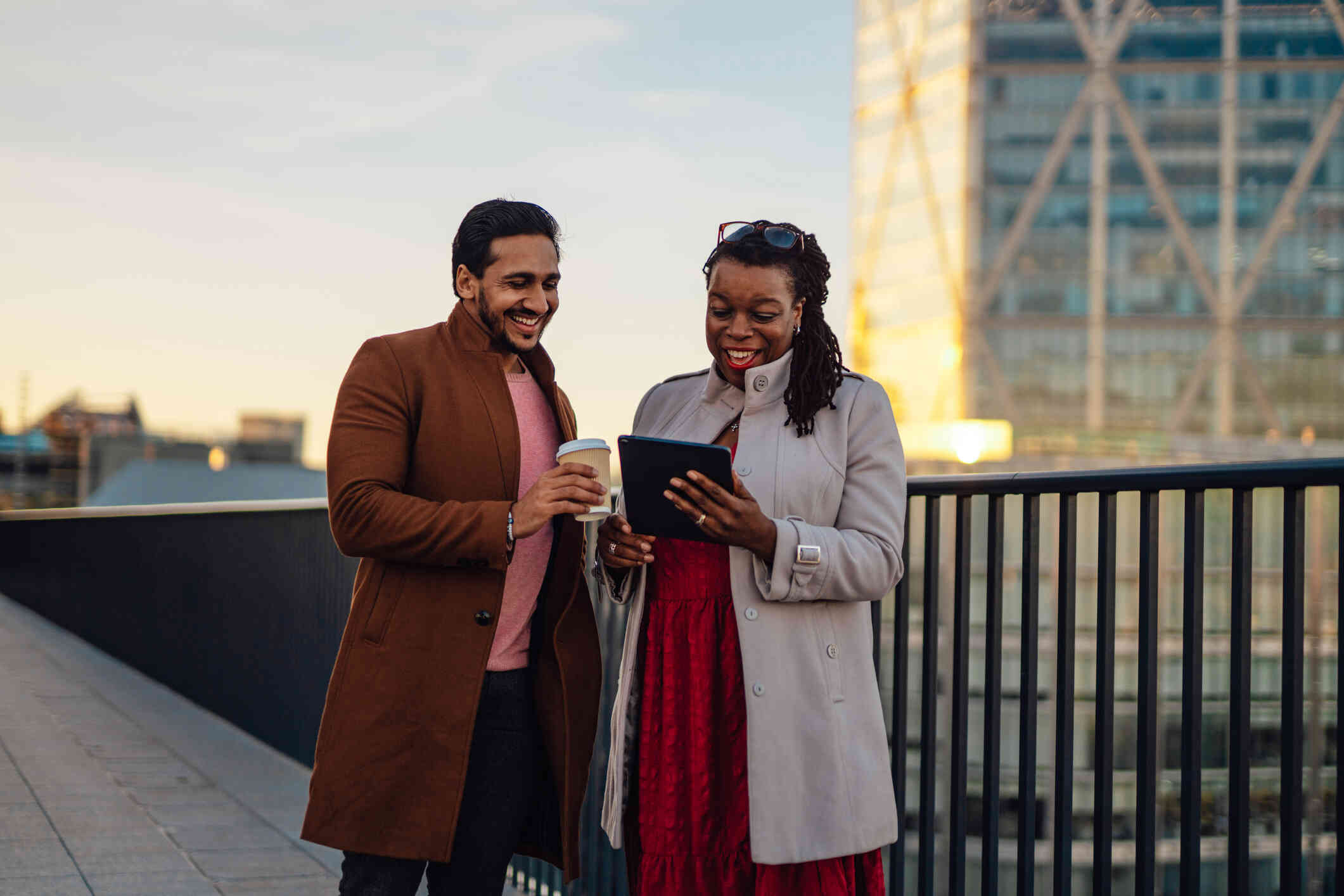 A man and woman in winter coats stand next to each other outside in the city and smile while looking at the womans tablet in her hand.