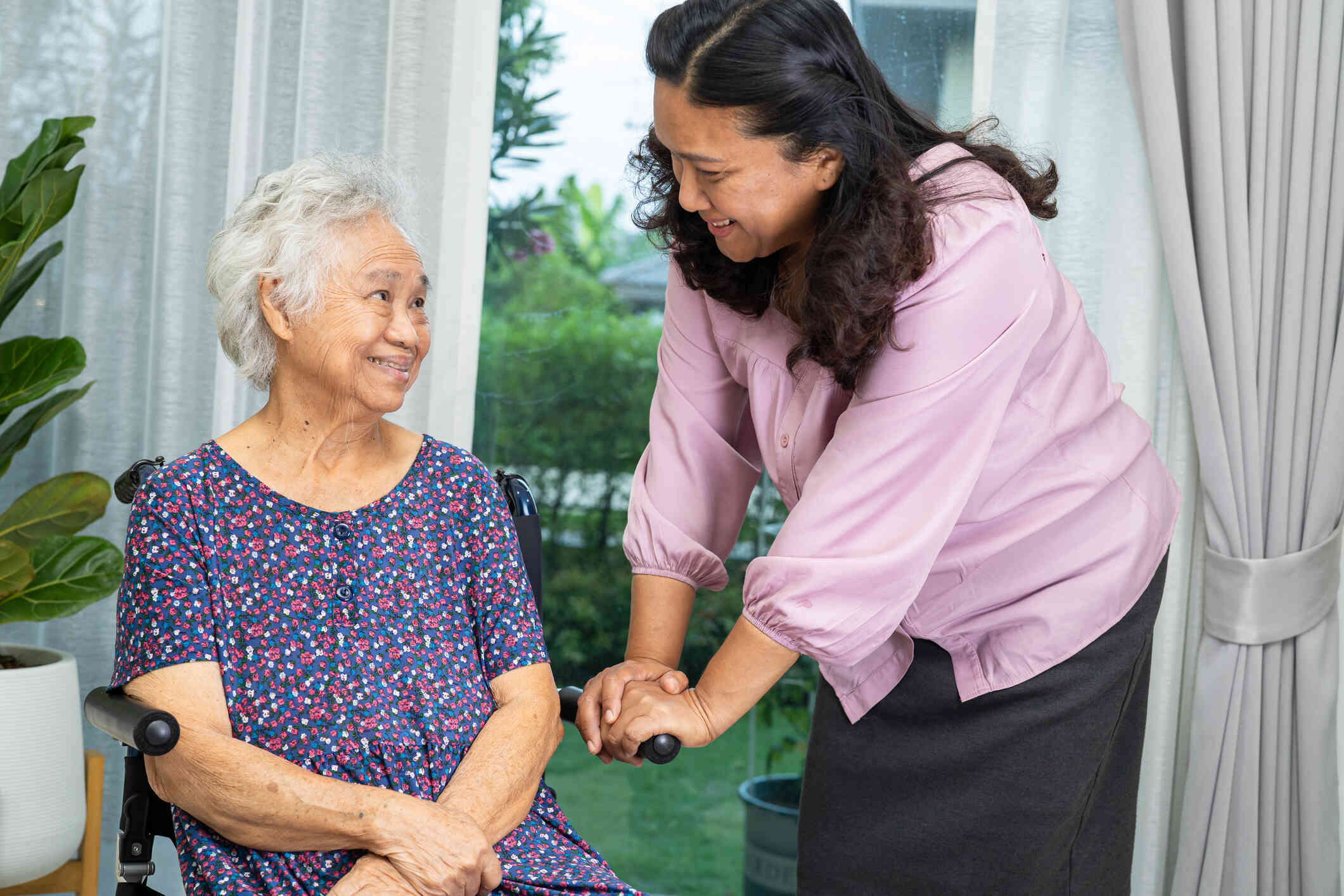 A woman in a pink shirt leans down to smile at an elderly female relative in a wheelchair.